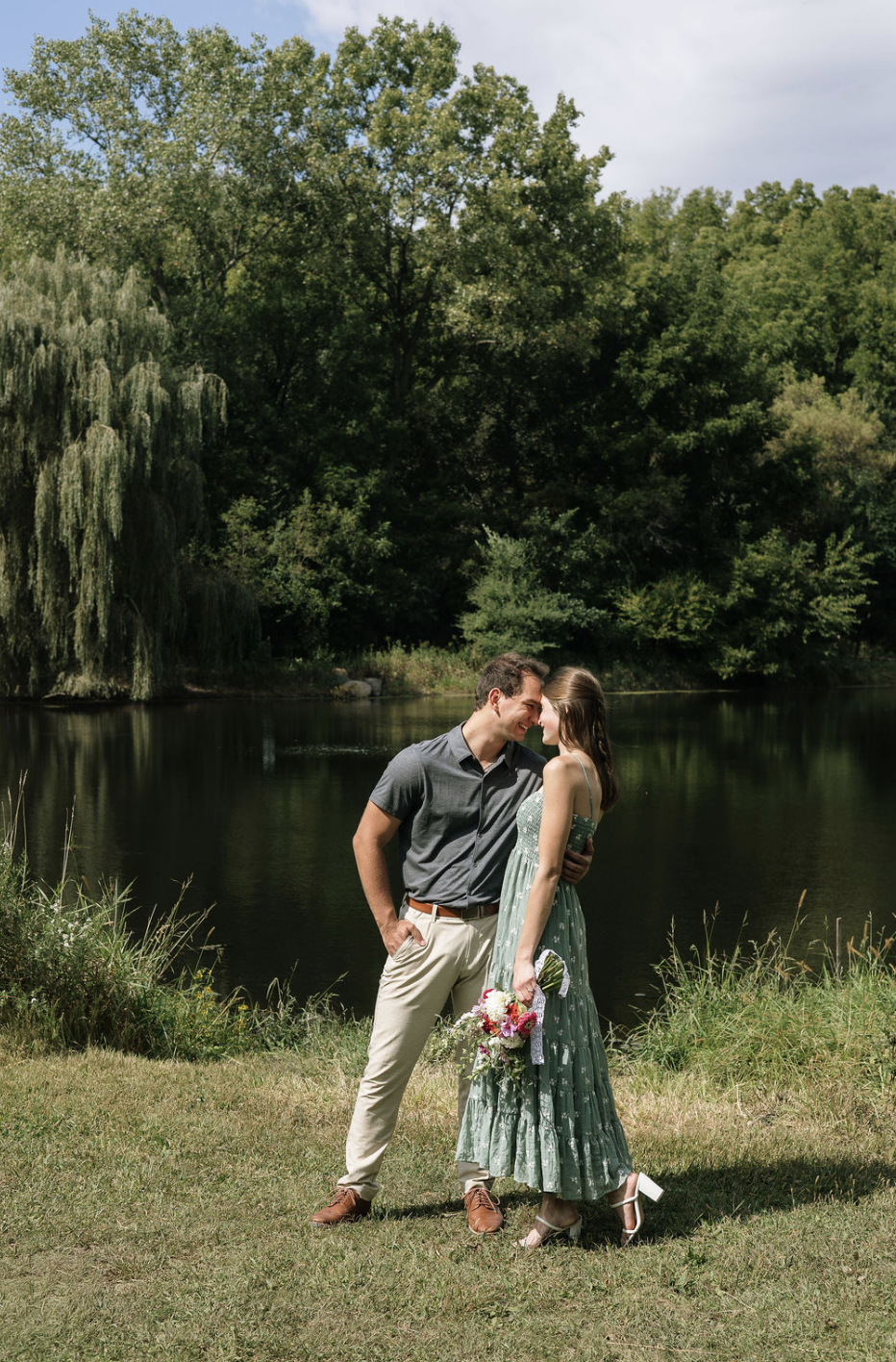 A couple standing close together by a lake, smiling and touching foreheads. The woman holds a bouquet of flowers, and the man has his hands in his pockets. They are outdoors with green trees and grass surrounding them.