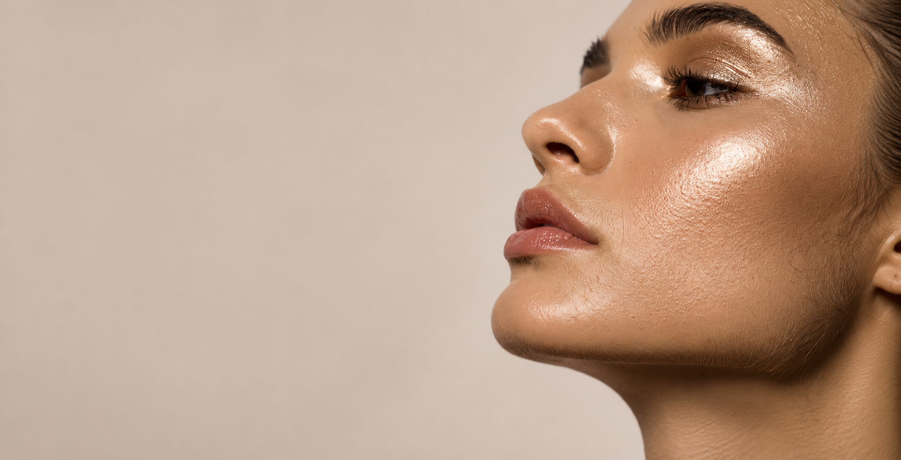Close-up of a woman's face in profile with makeup and glowing skin against a neutral background.