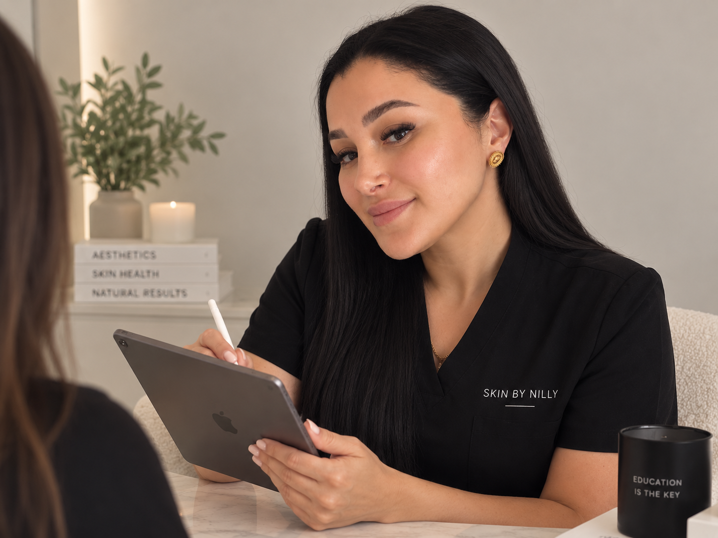 A woman with black hair and gold earrings sitting at a table, writing on a tablet with a stylus, talking to another person. She is wearing a black uniform with 'Skin by Nilly' embroidered on it. In the background, there is a white sign with black text reading 'AESTHETICS, SKIN HEALTH, NATURAL RESULTS', and a potted plant and candle.
