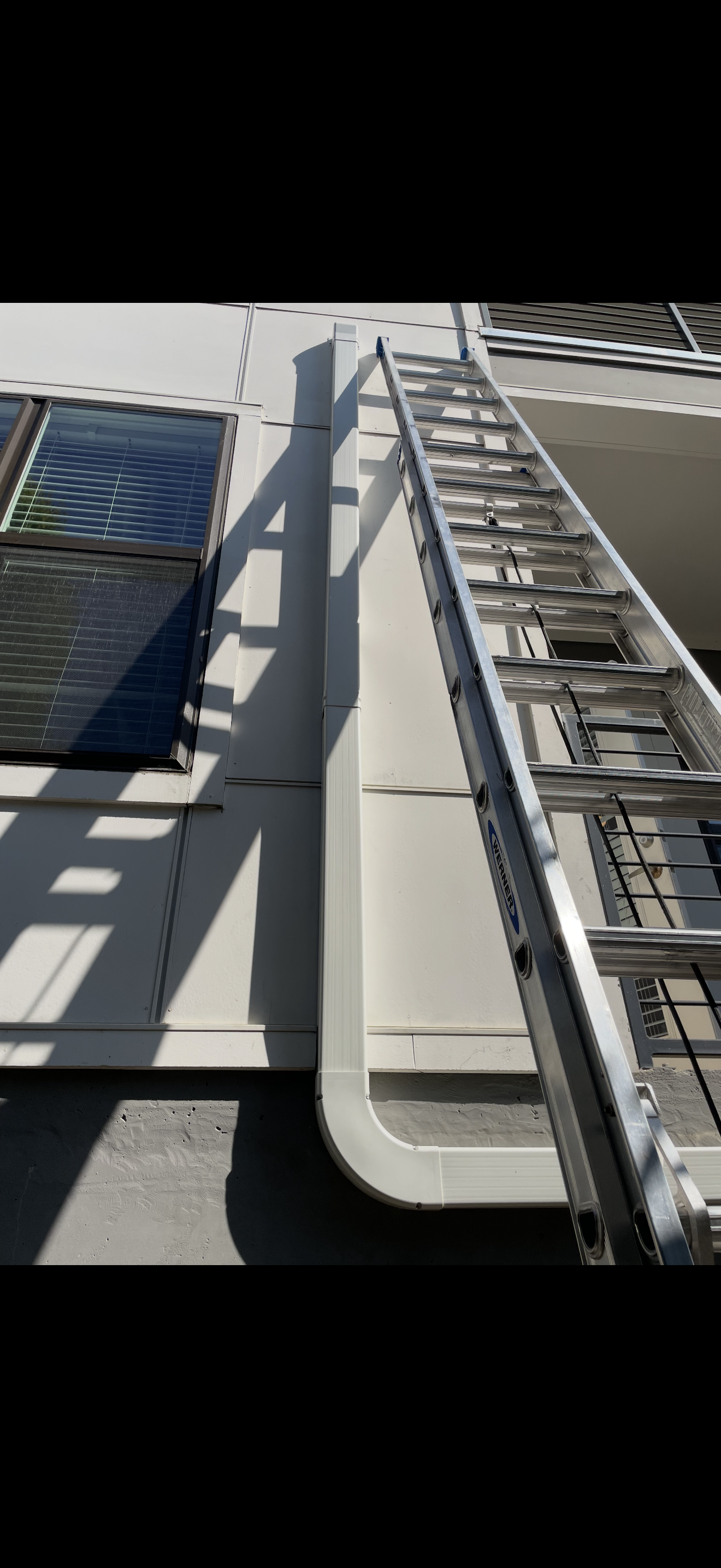 An exterior view of a building showing a window with blinds, a downspout, and an extension ladder leaning against the wall.