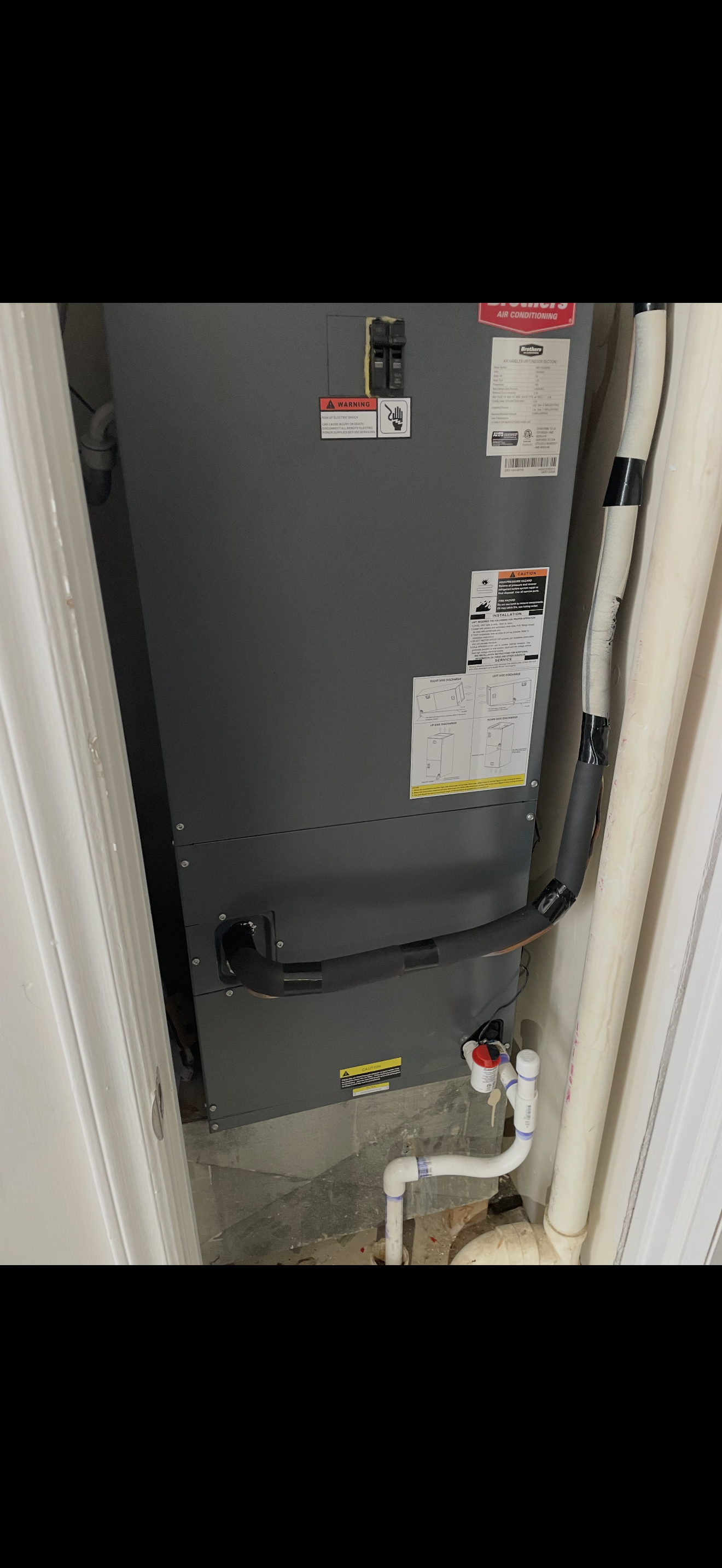 A gray HVAC furnace unit with various warning labels, pipes, and electrical connections installed in a utility closet.