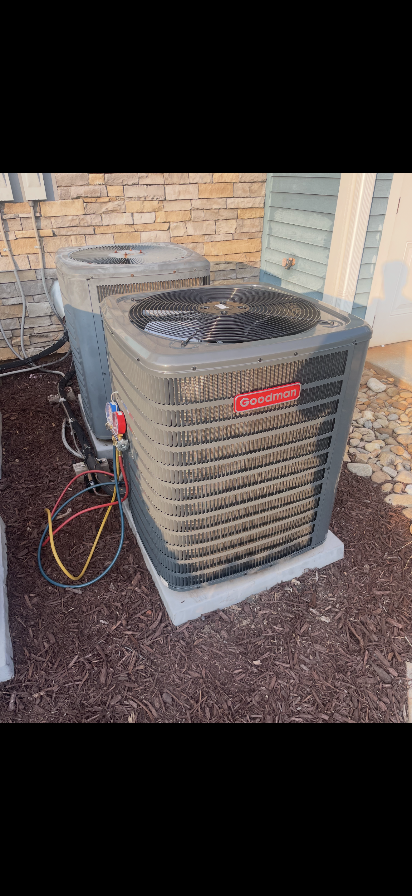 Outside air conditioning unit with a Goodman logo on a concrete pad, connected with red, yellow, and blue wires, next to a house wall with brick and siding, and surrounded by mulch and rocks.