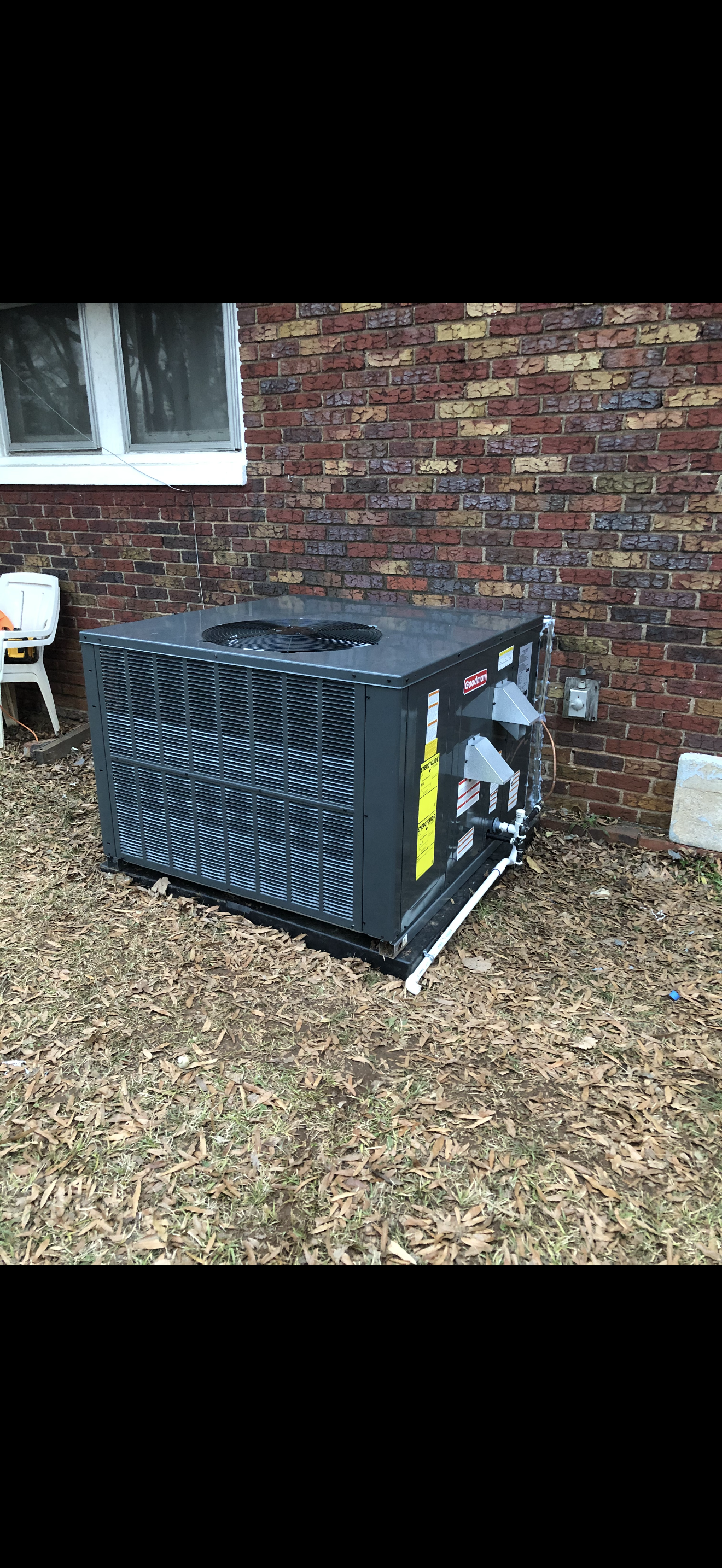 Outdoor heat pump unit installed against a brick wall with a window above, on a ground covered with dry leaves and grass.