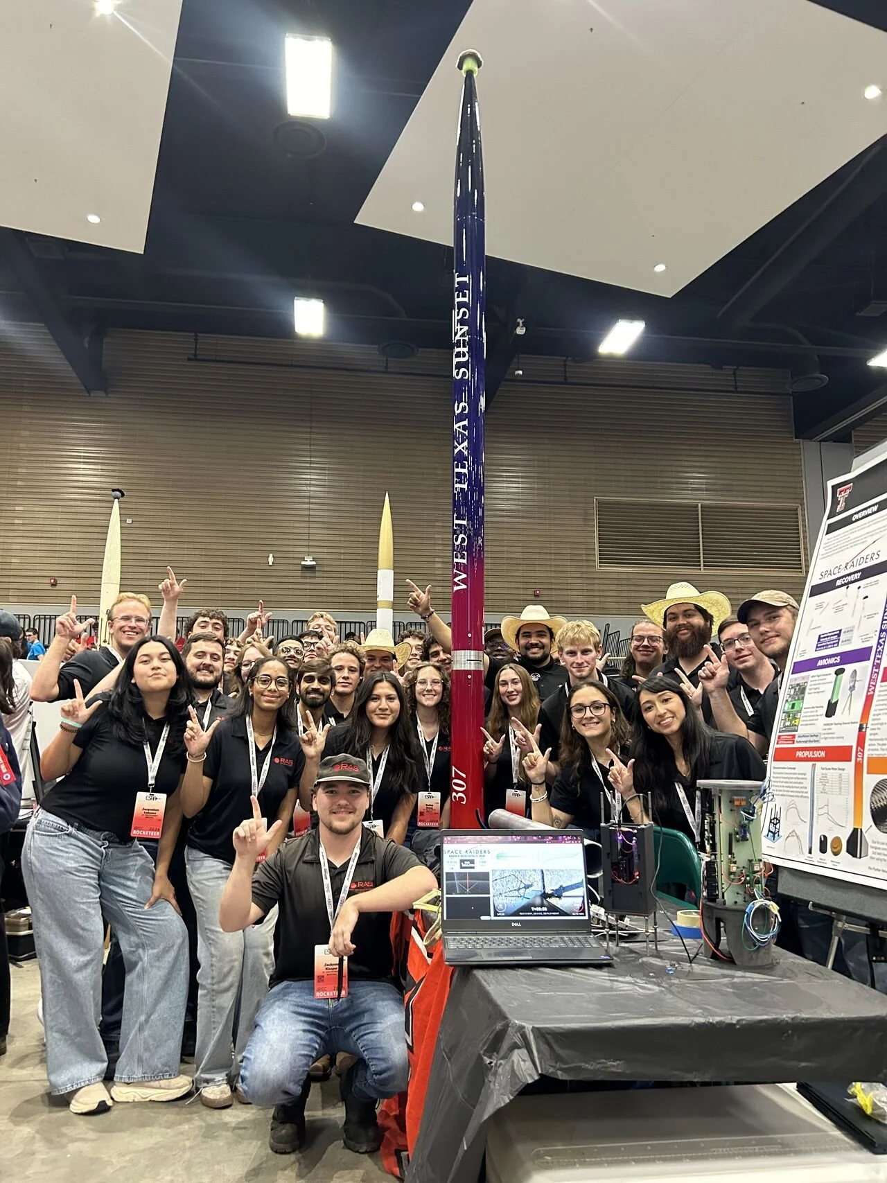 Group of students and team members at a science or engineering competition, posing around a tall rocket with a crowd of people in the background.