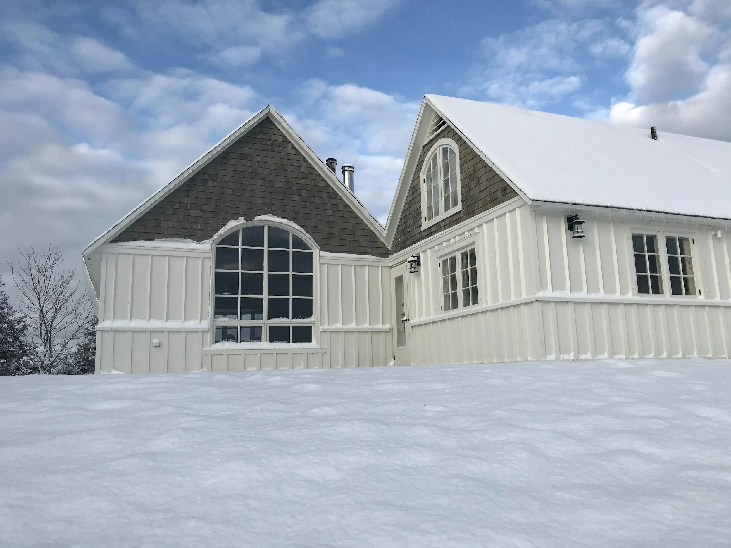 A house with a gabled roof covered in snow, large arched window and white siding, surrounded by snow, under a partly cloudy sky.