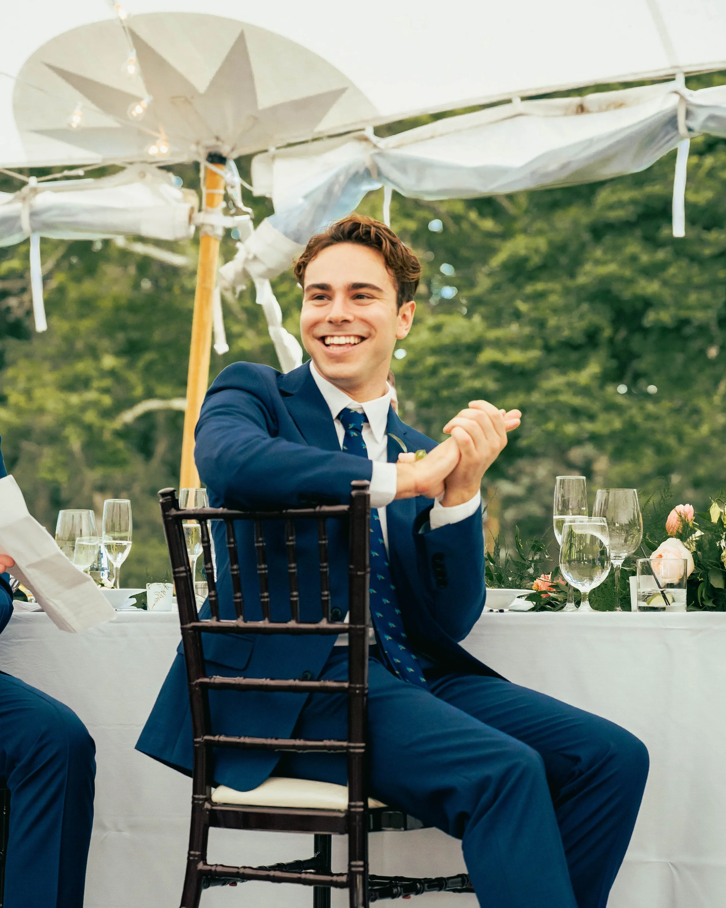 Man in a blue suit smiling and gesturing with his hands at an outdoor event with tables, glasses, and decorative umbrellas.