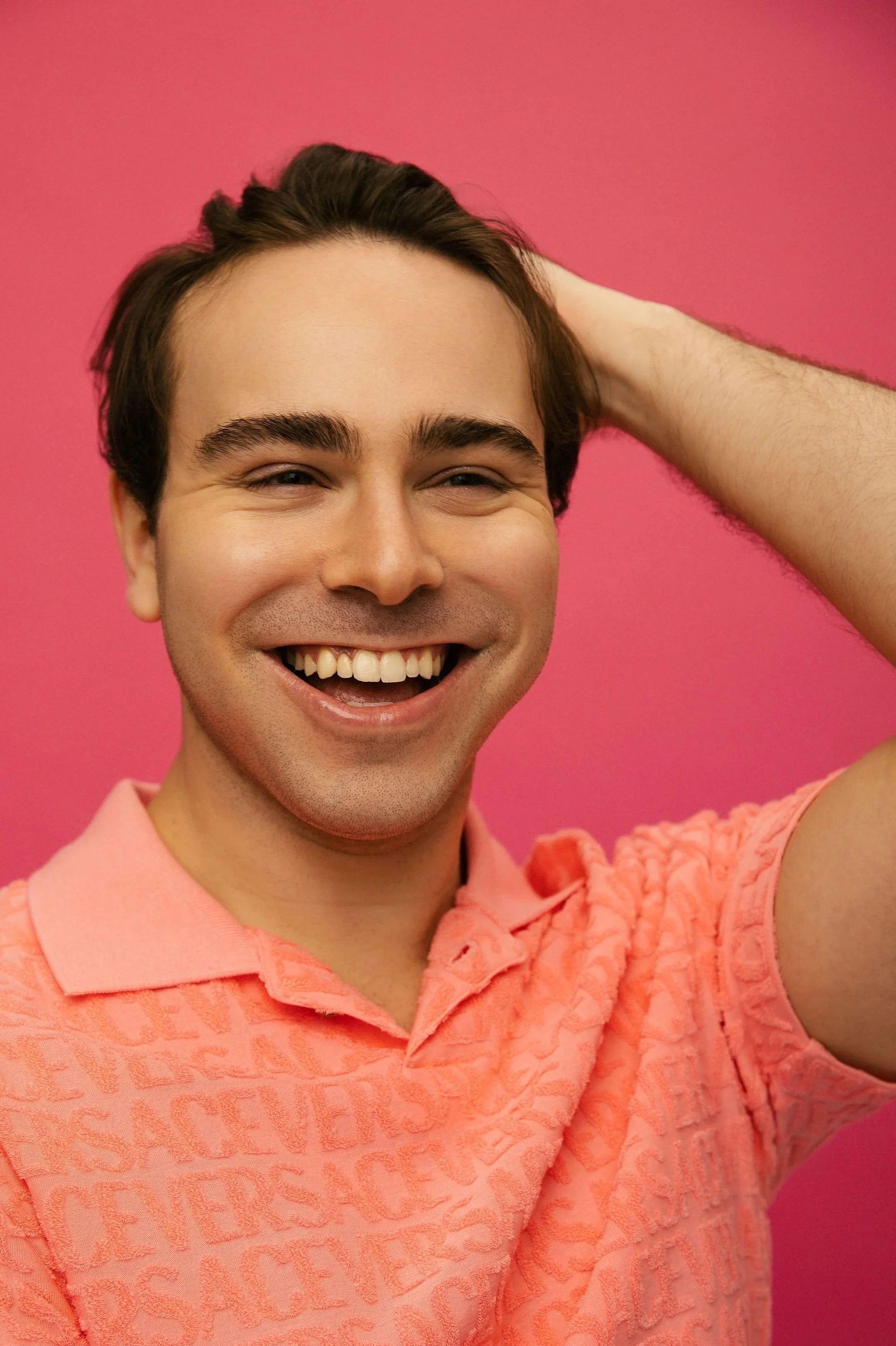 A smiling young man with dark brown hair and eyebrows, wearing a pink polo shirt, against a solid pink background.