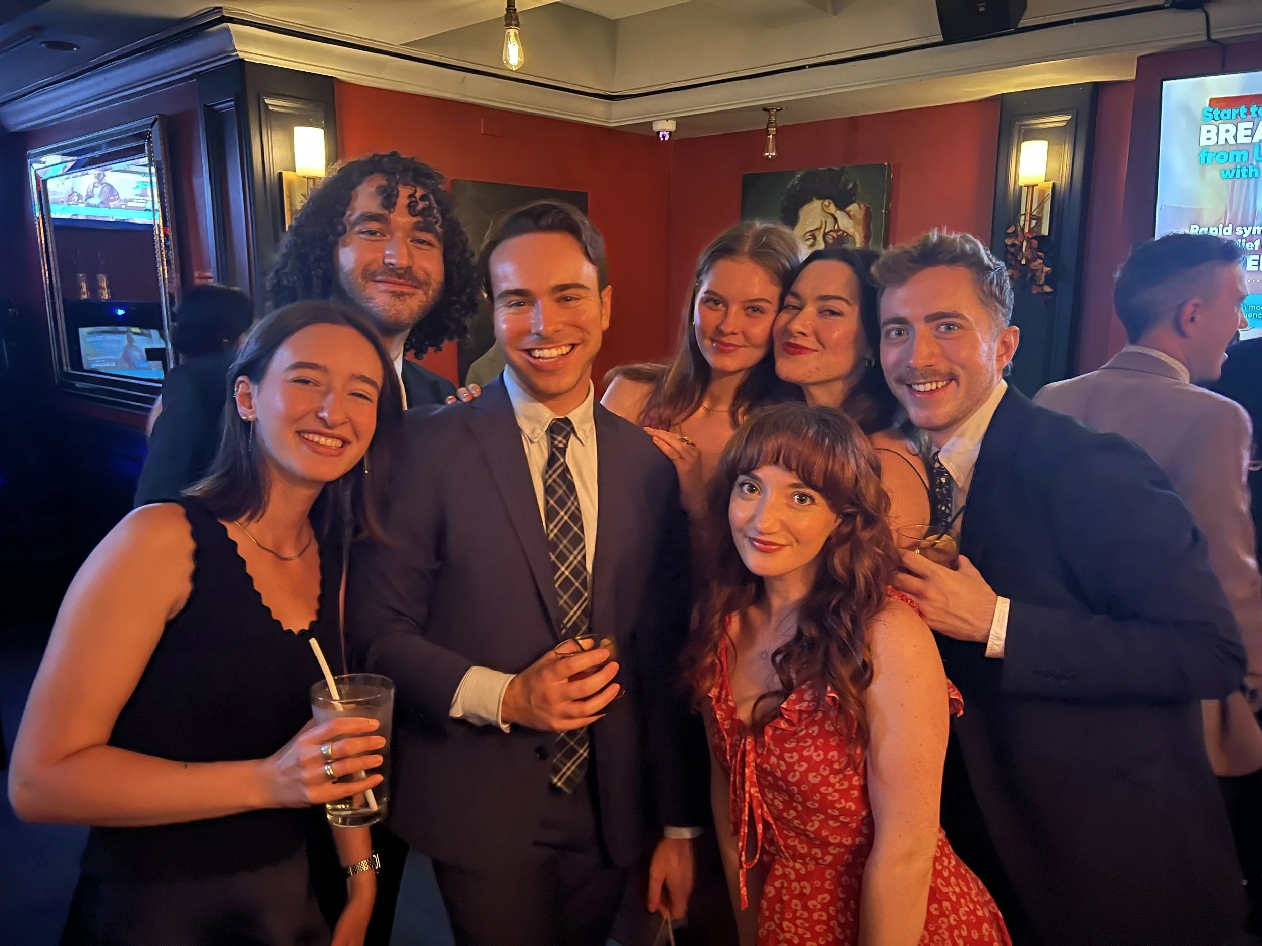 Group of six young adults, dressed up, smiling, and enjoying drinks at a party or social gathering in a dimly lit indoor venue.