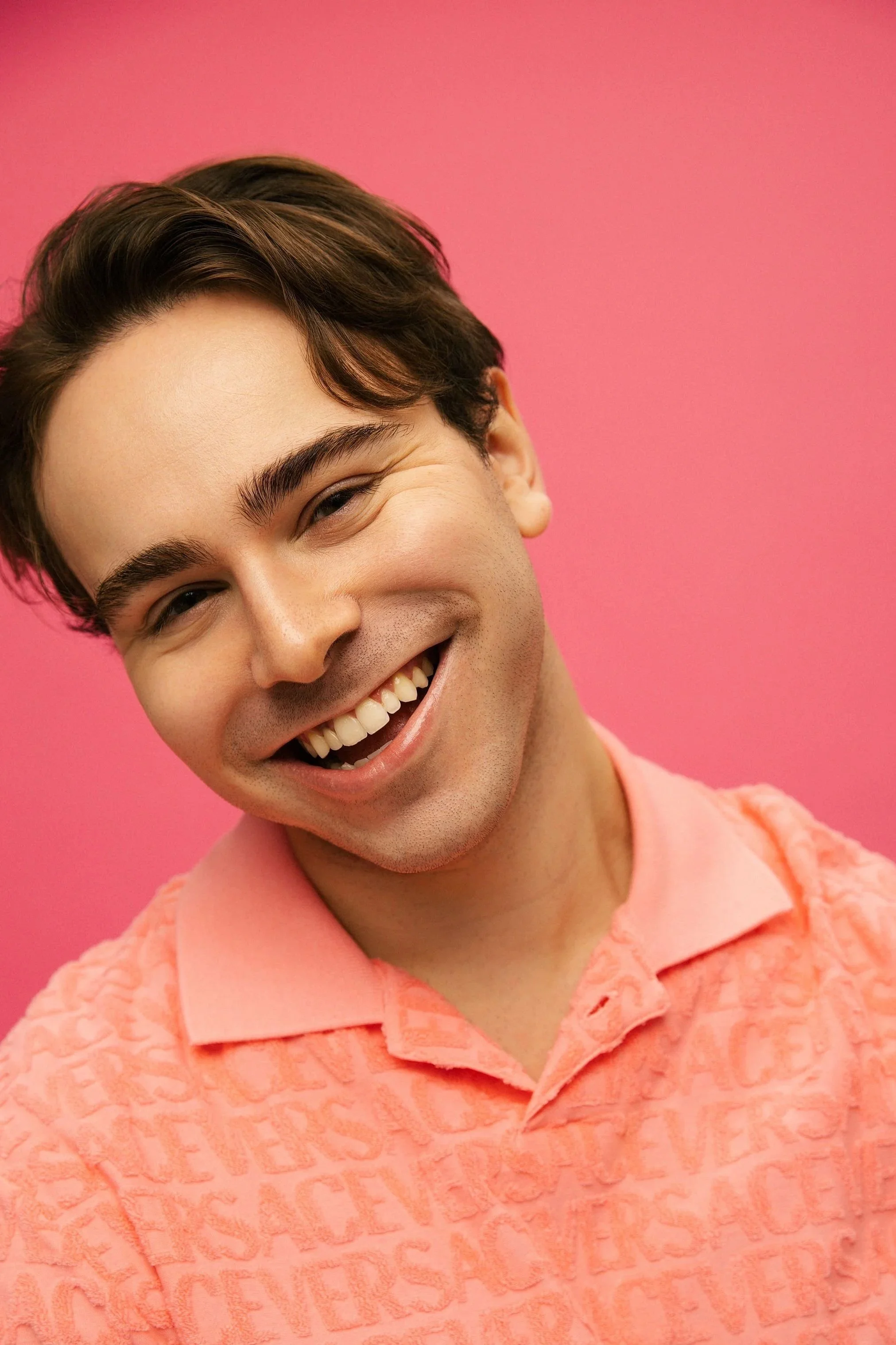 Close-up of a smiling young man with dark hair, wearing a pink textured shirt, against a pink background.