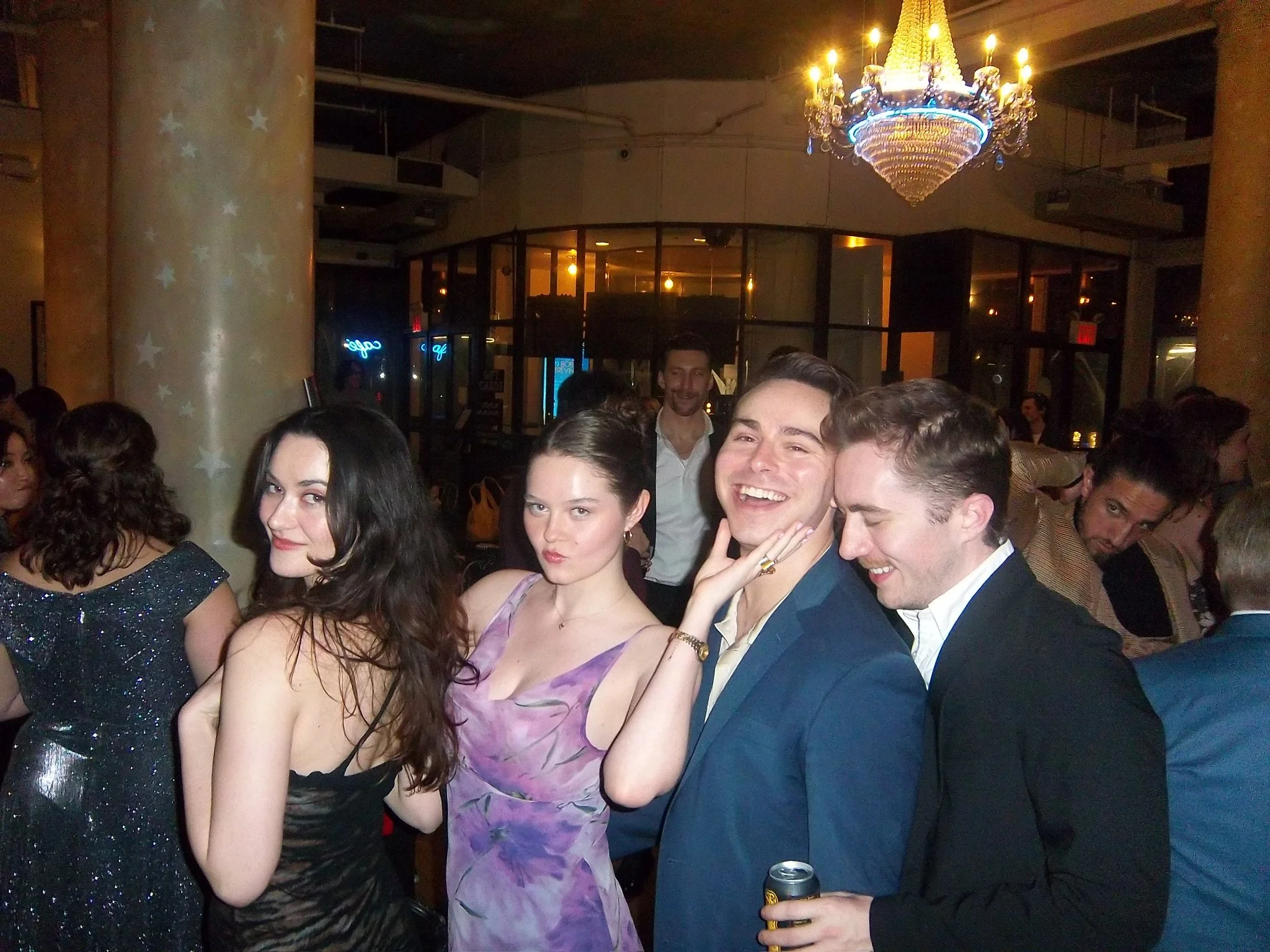 A group of young adults dressed in formal attire socializing at an indoor event with a chandelier hanging from the ceiling, and large glass windows in the background.