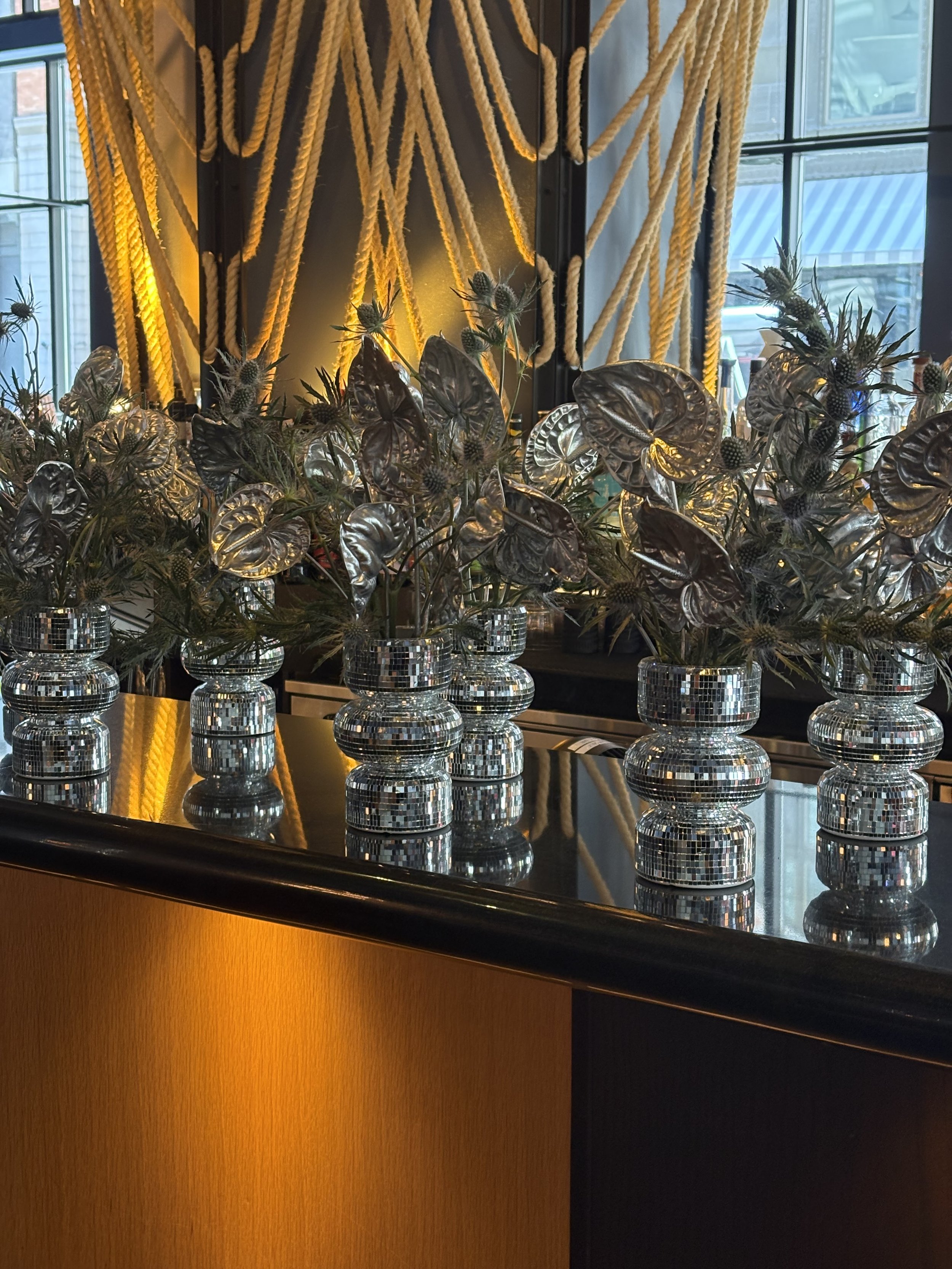 Interior decor with multiple glass vases holding silver-colored plants, set on a reflective black surface. Behind, there are yellow ropes arranged decoratively on a dark wall with backlighting, next to large windows showing an urban exterior.