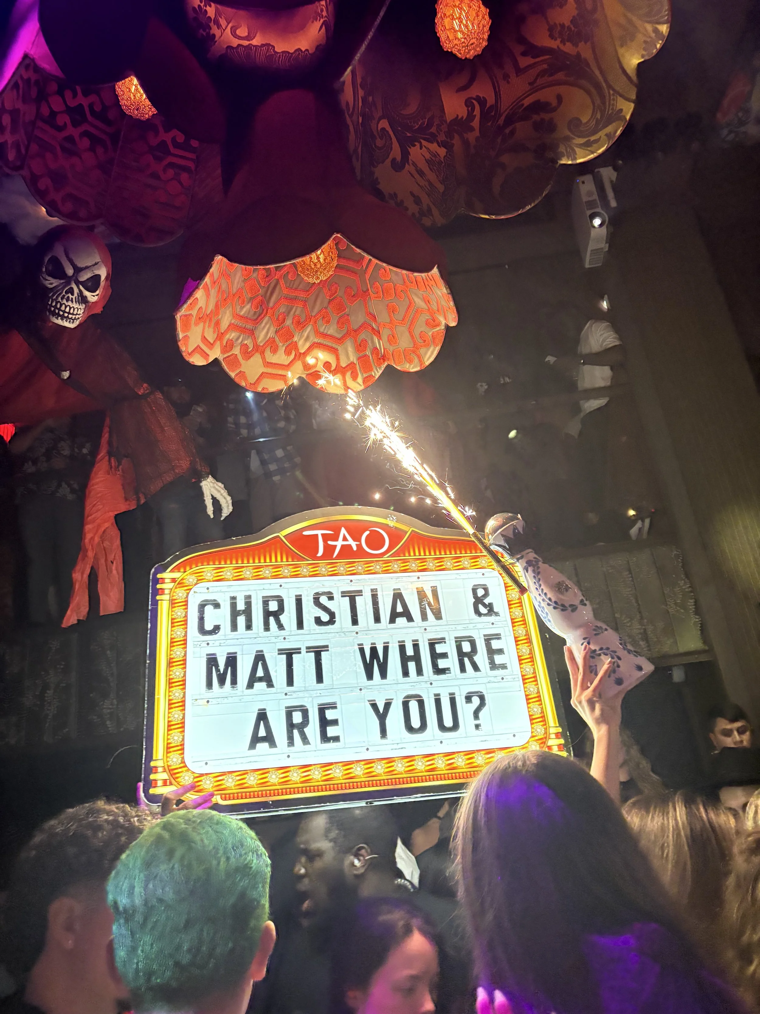 Crowd at a party or celebration with colorful decorations, a sign reading 'Christian & Matt Where Are You?', a skull mask, balloons, and a sparkler firework.