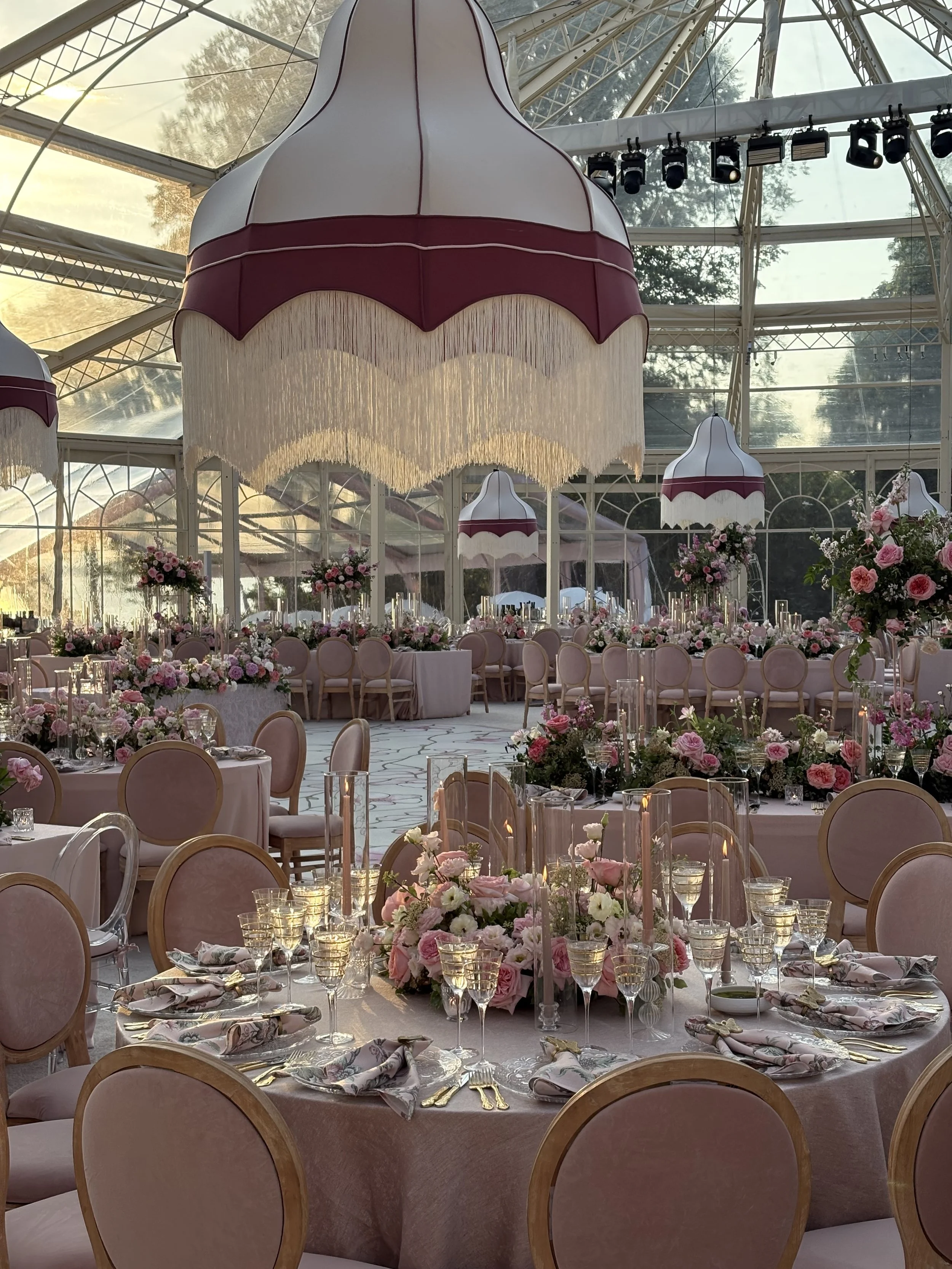 Elegant indoor wedding reception setup with pink and white floral arrangements, pink cushioned chairs, and large pink flower-shaped chandeliers hanging from the glass ceiling.