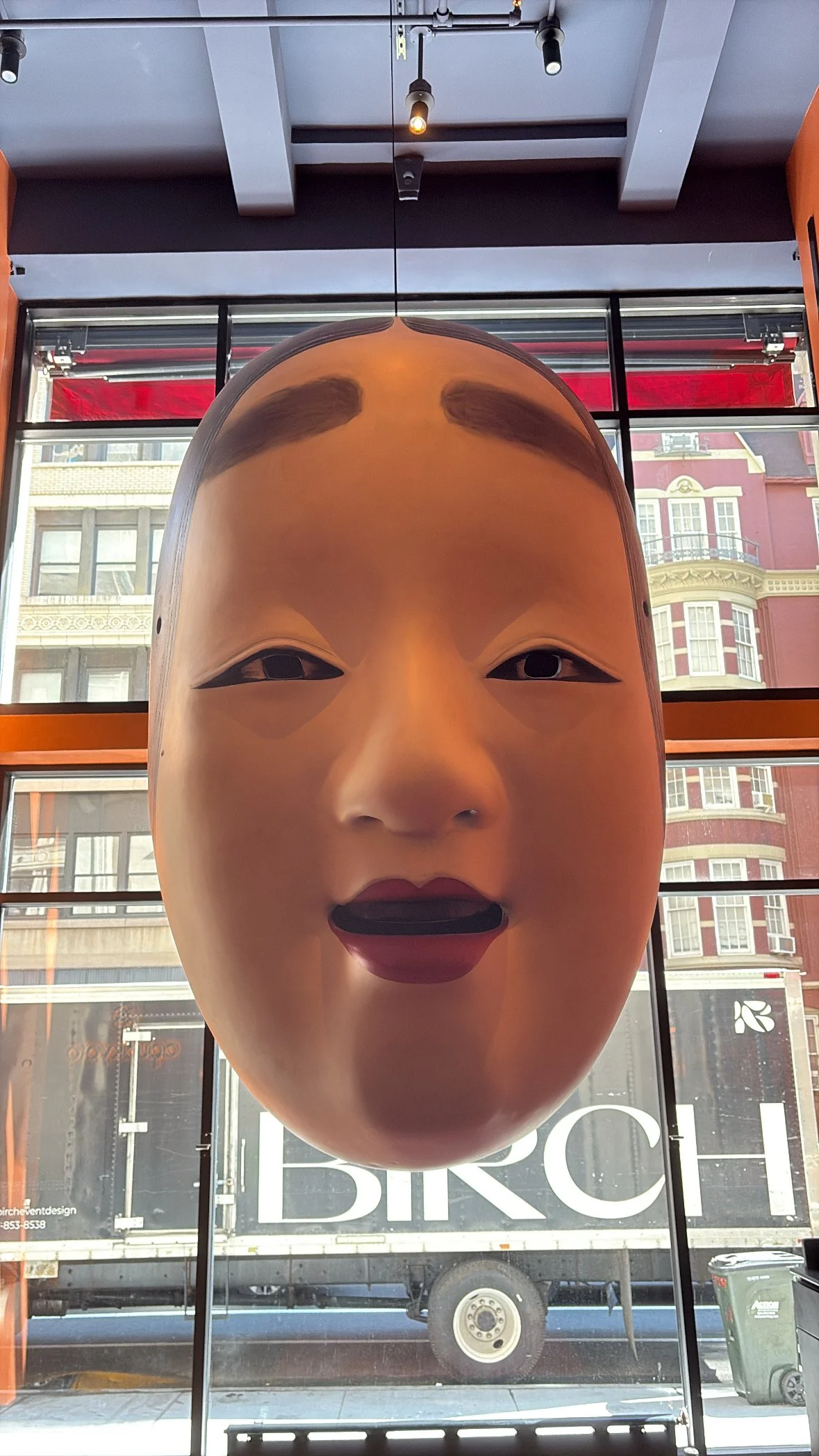 Large Asian-style face mask with exaggerated facial features, including arched eyebrows, slanted eyes, a small nose, and full lips, displayed indoors with a window showing city buildings and a truck outside.