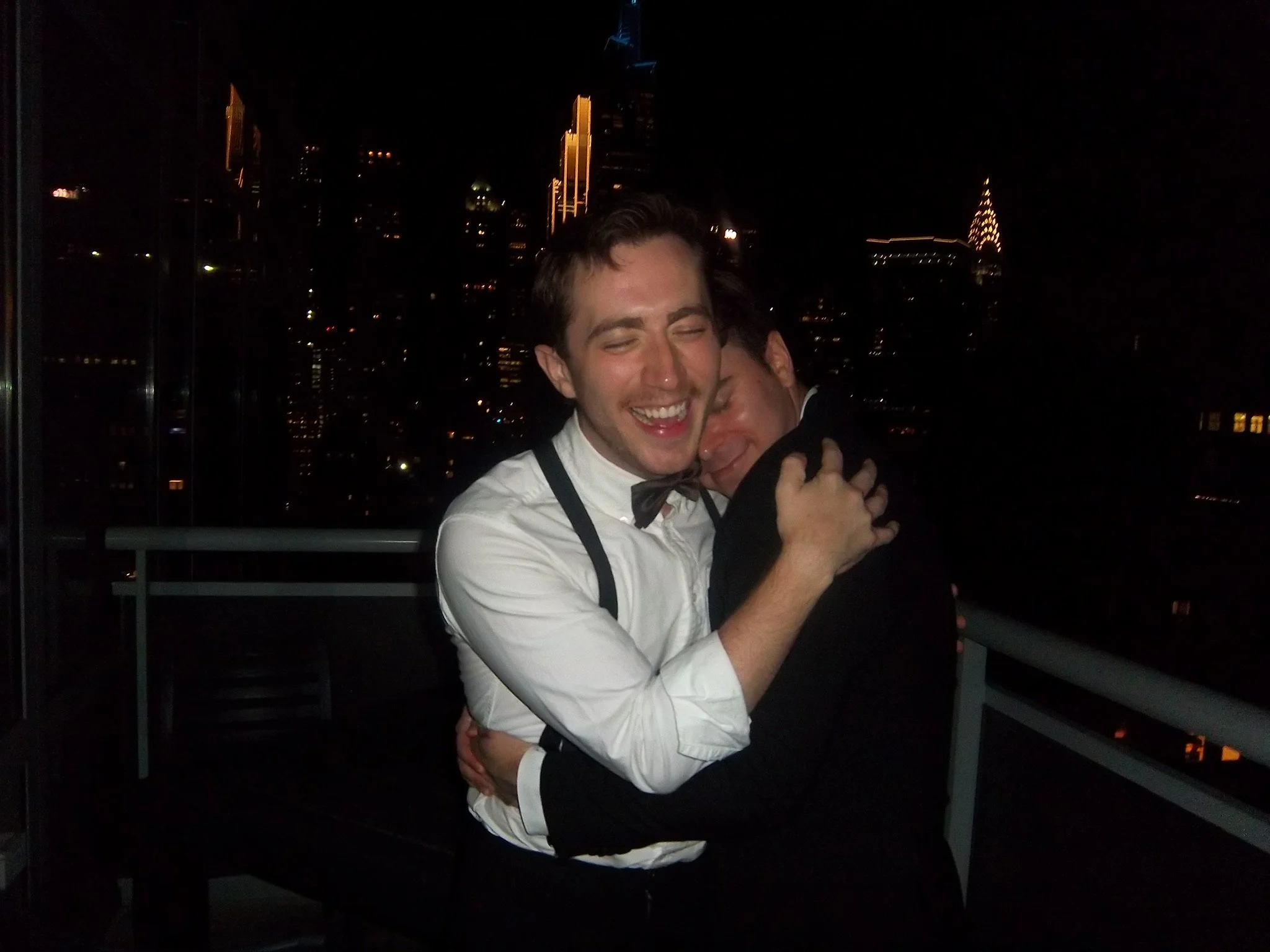 Two people dressed in formal attire embracing on a balcony at night with a city skyline, including tall illuminated buildings, in the background.