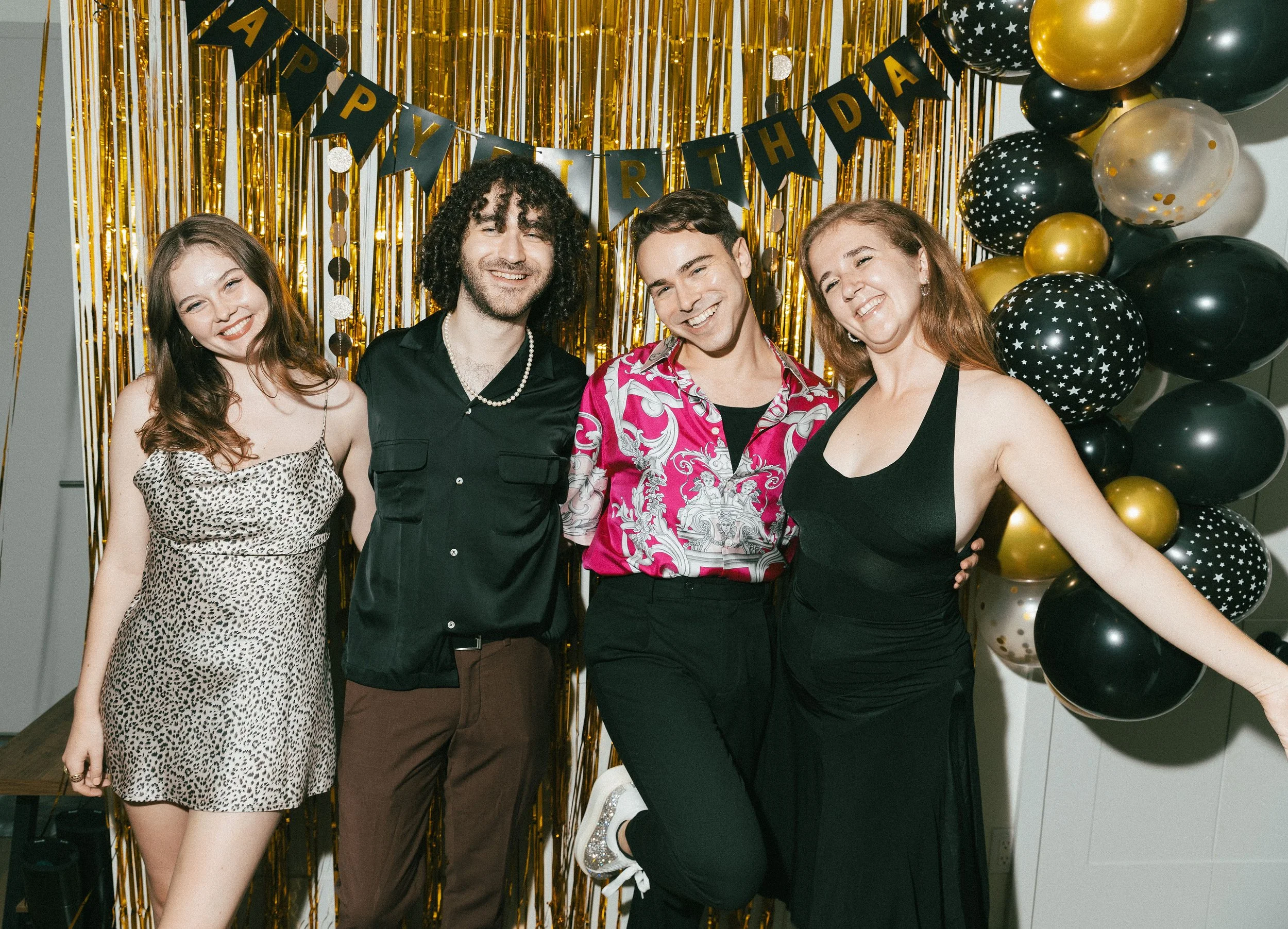Group of four friends smiling and posing together at a birthday party with black, gold, and star-patterned balloons and a "Happy Birthday" banner in the background.