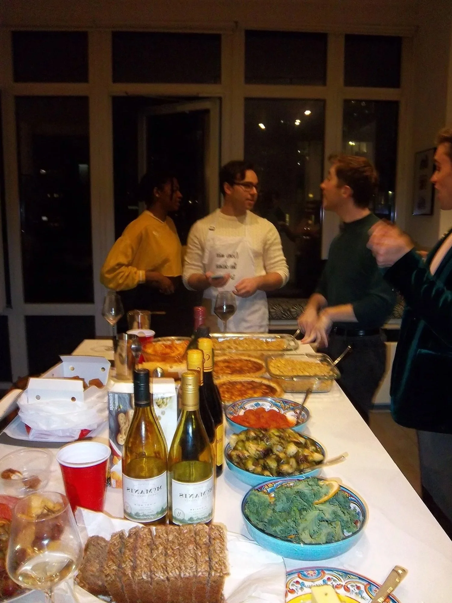 A group of people in a kitchen or dining area preparing or serving food, with plates of pizza, salads, and bottles of wine on the table.