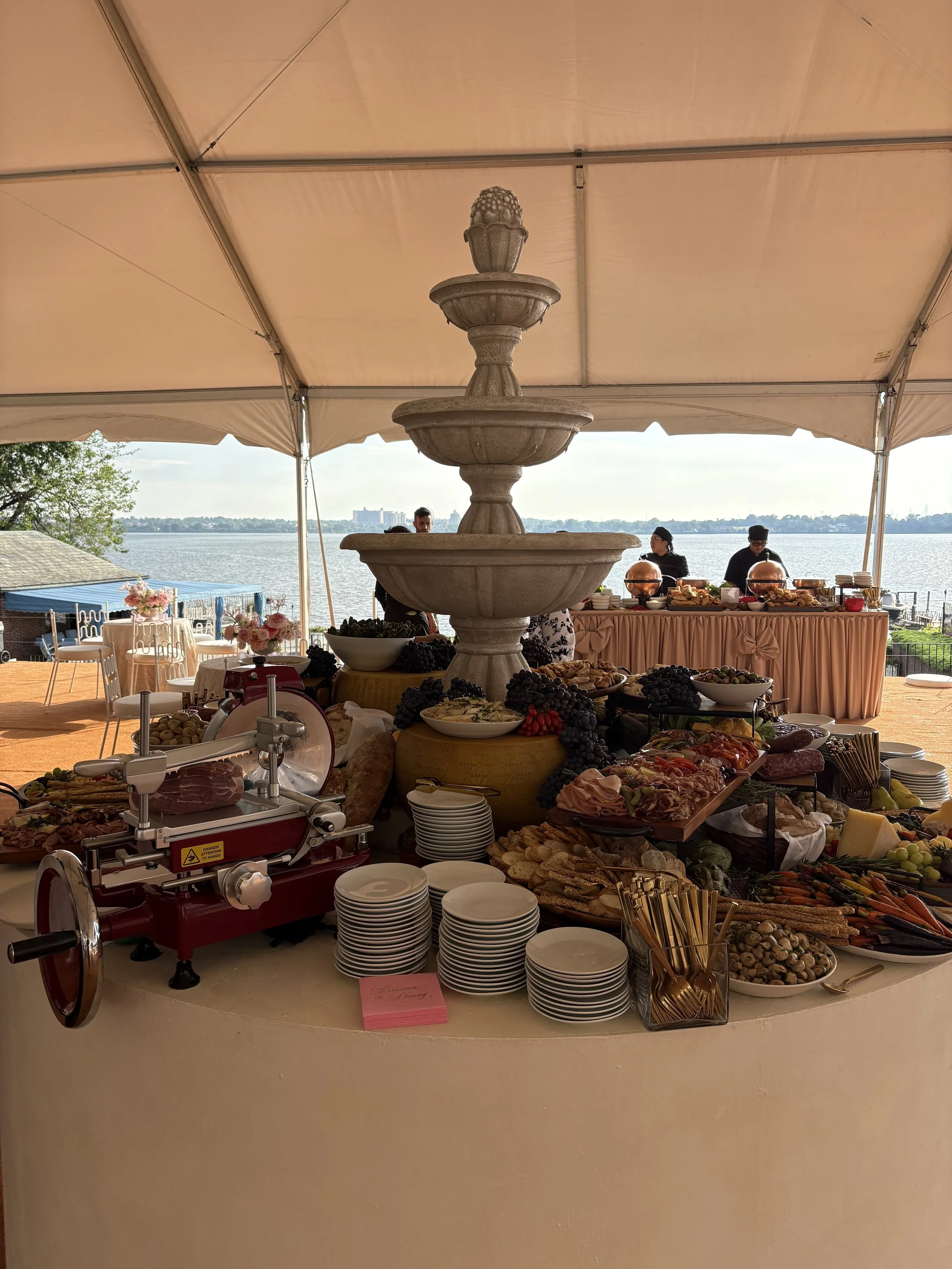 A buffet table with various foods near a large fountain with water, overlooking a body of water and distant buildings, under a canopy at an outdoor event.