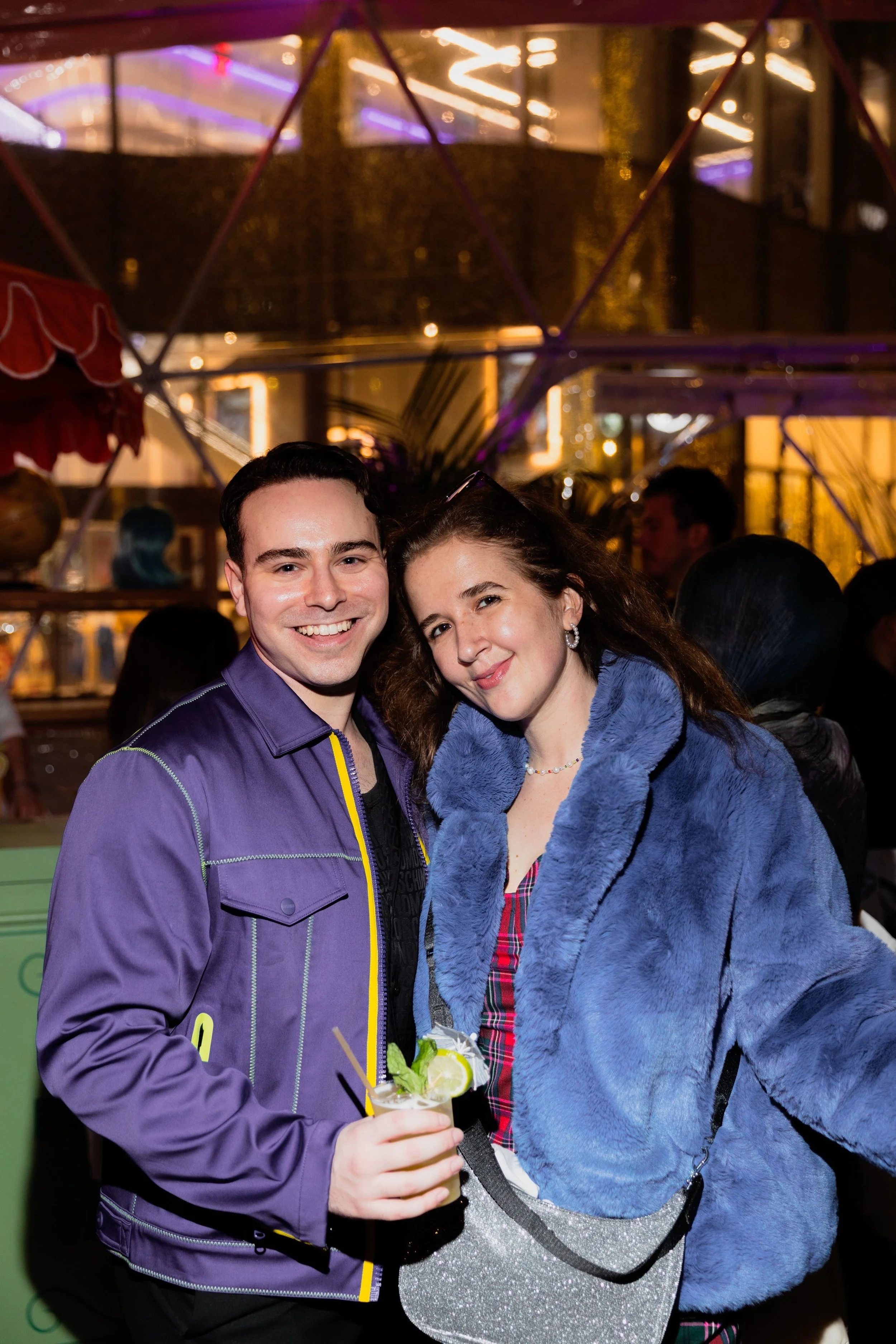 A smiling young man and woman posing together at night at a social event, with people and decorative lights in the background. The man is holding a cocktail drink garnished with lime and mint.