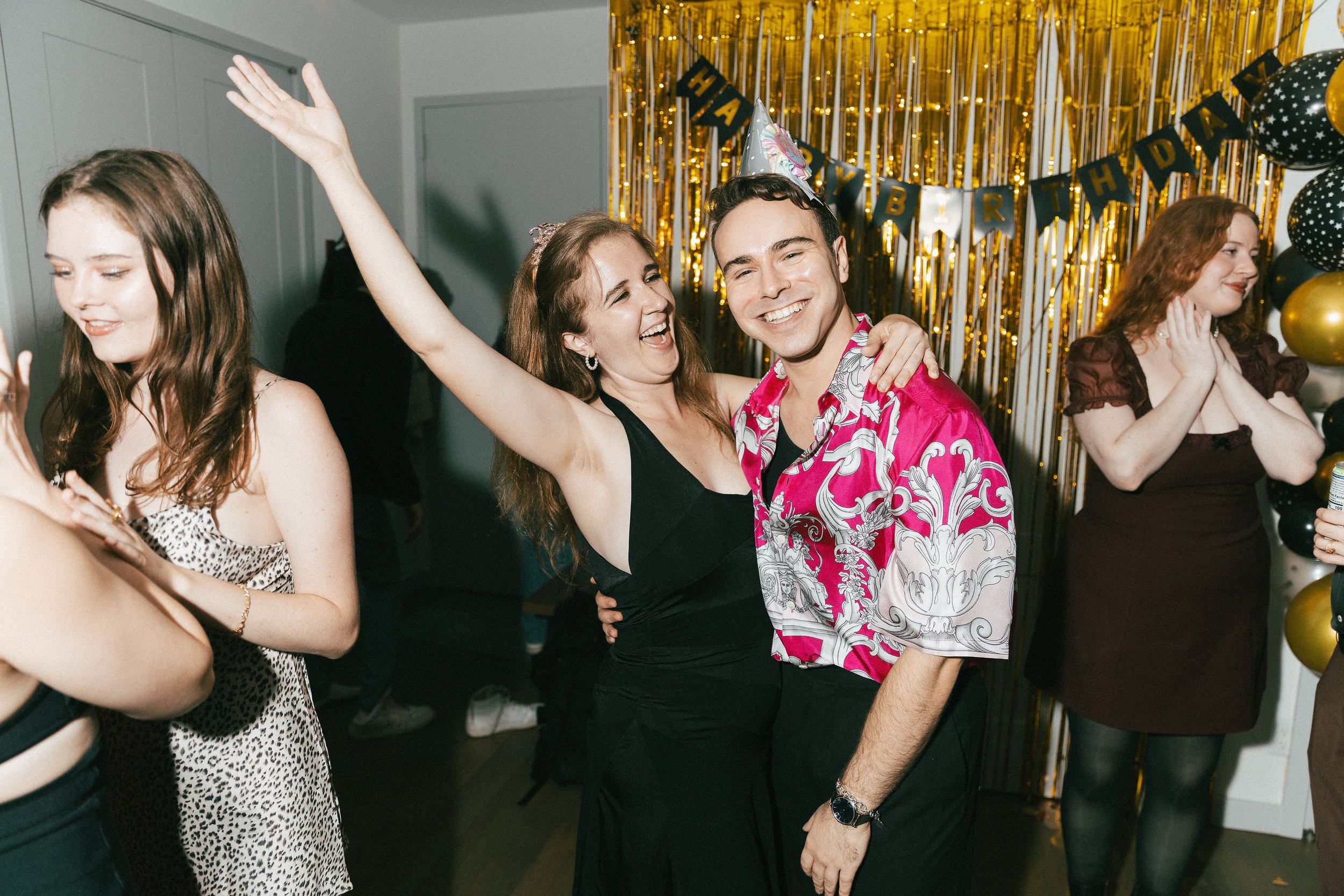 Group of people celebrating at a birthday party with gold and black decorations, some wearing birthday hats.
