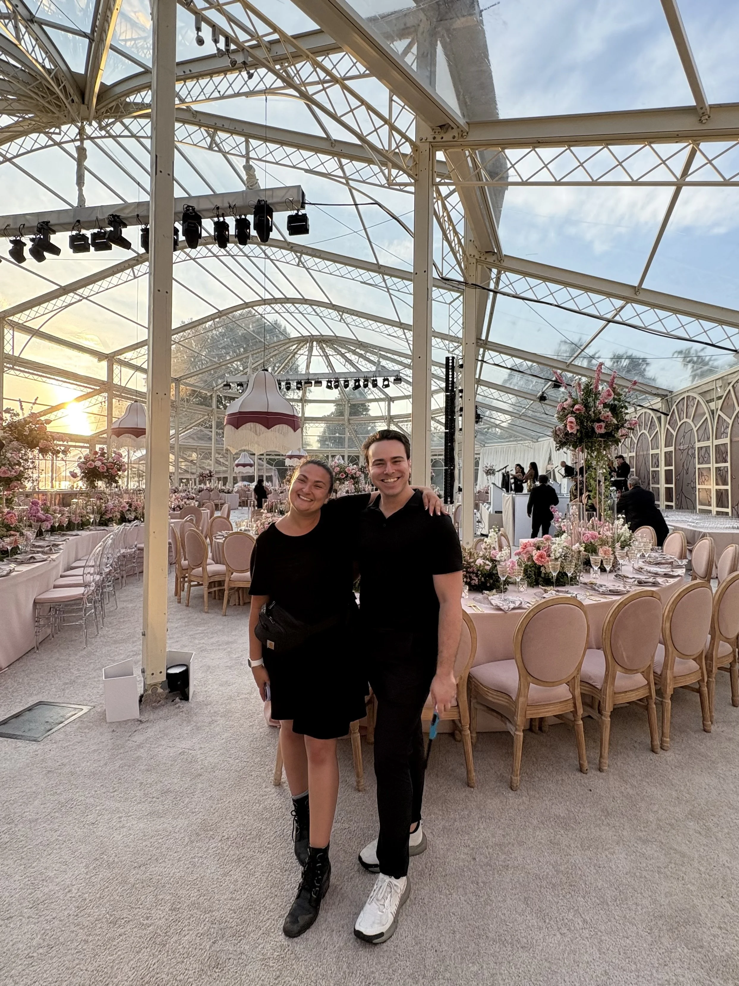 A smiling couple stands close together inside a bright, elegant event venue with decorated tables, floral centerpieces, and a glass ceiling.