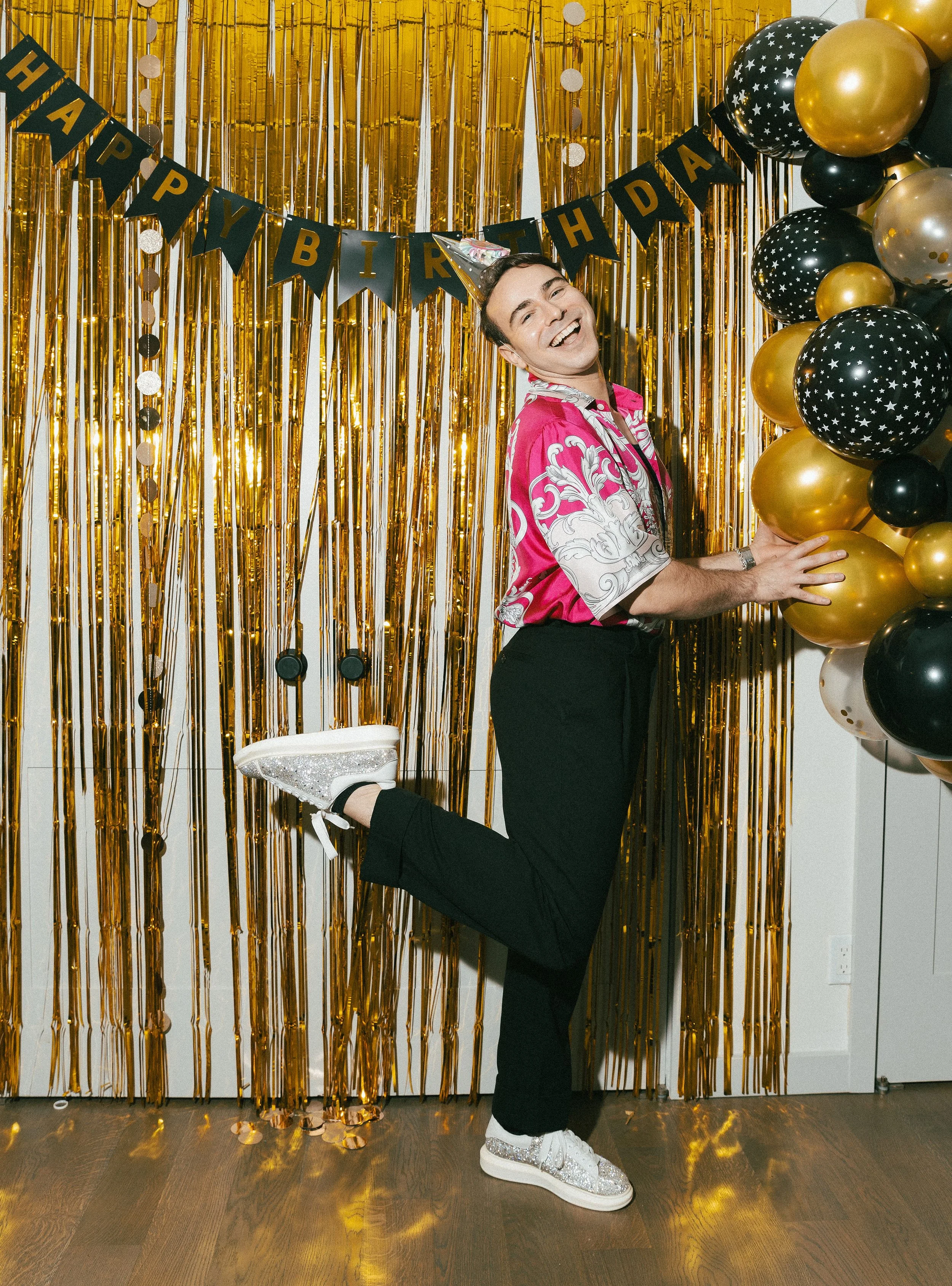 A person celebrating a birthday party standing on one leg with the other leg bent behind, wearing a party hat and a vibrant pink shirt, against a gold and black balloon backdrop with a 'Happy Birthday' banner.