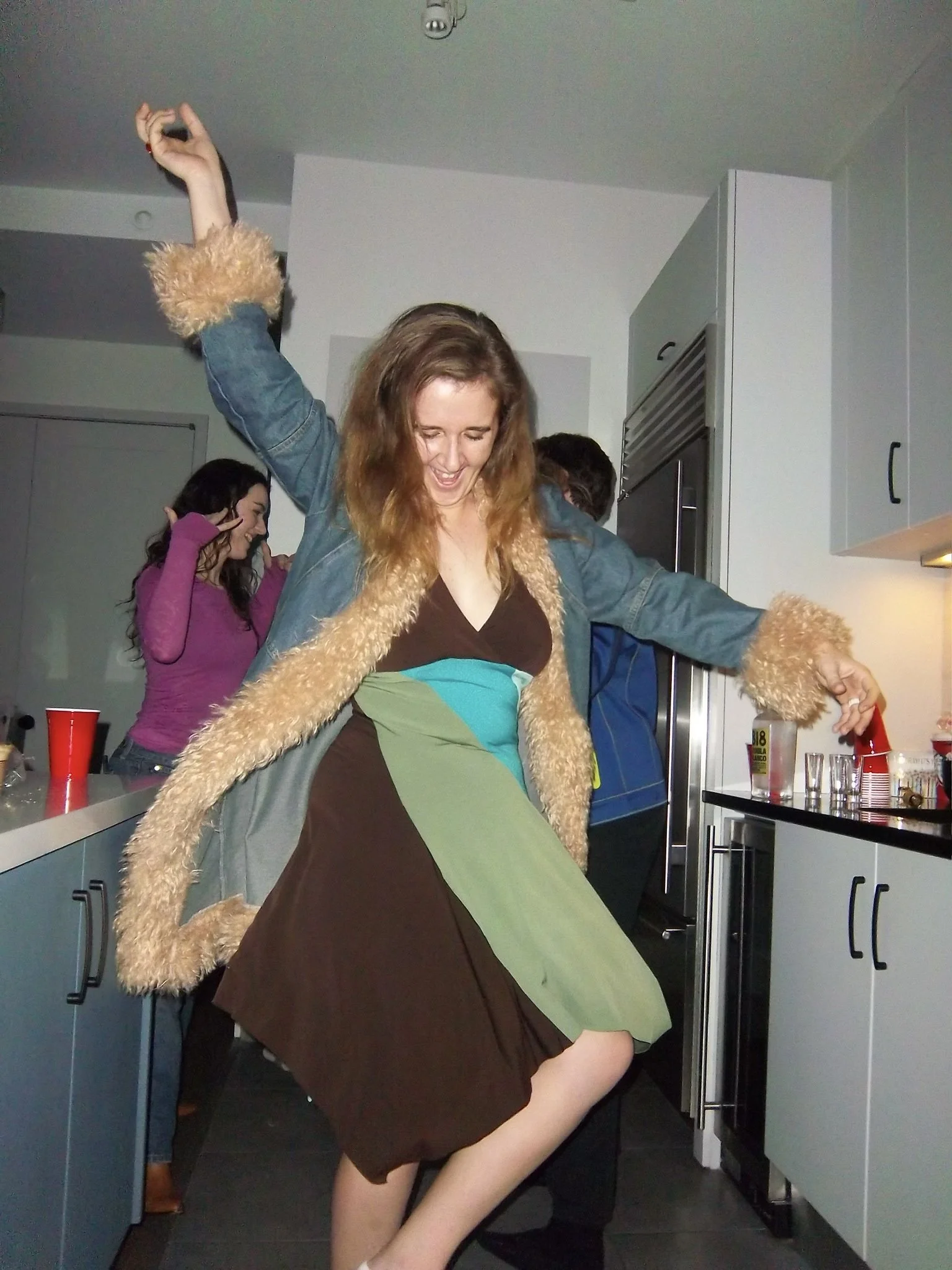 A young woman with long red hair dancing at a party. She is wearing a multicolored dress and a denim jacket with fur trim. She appears to be enjoying herself with her eyes closed and a smile on her face.