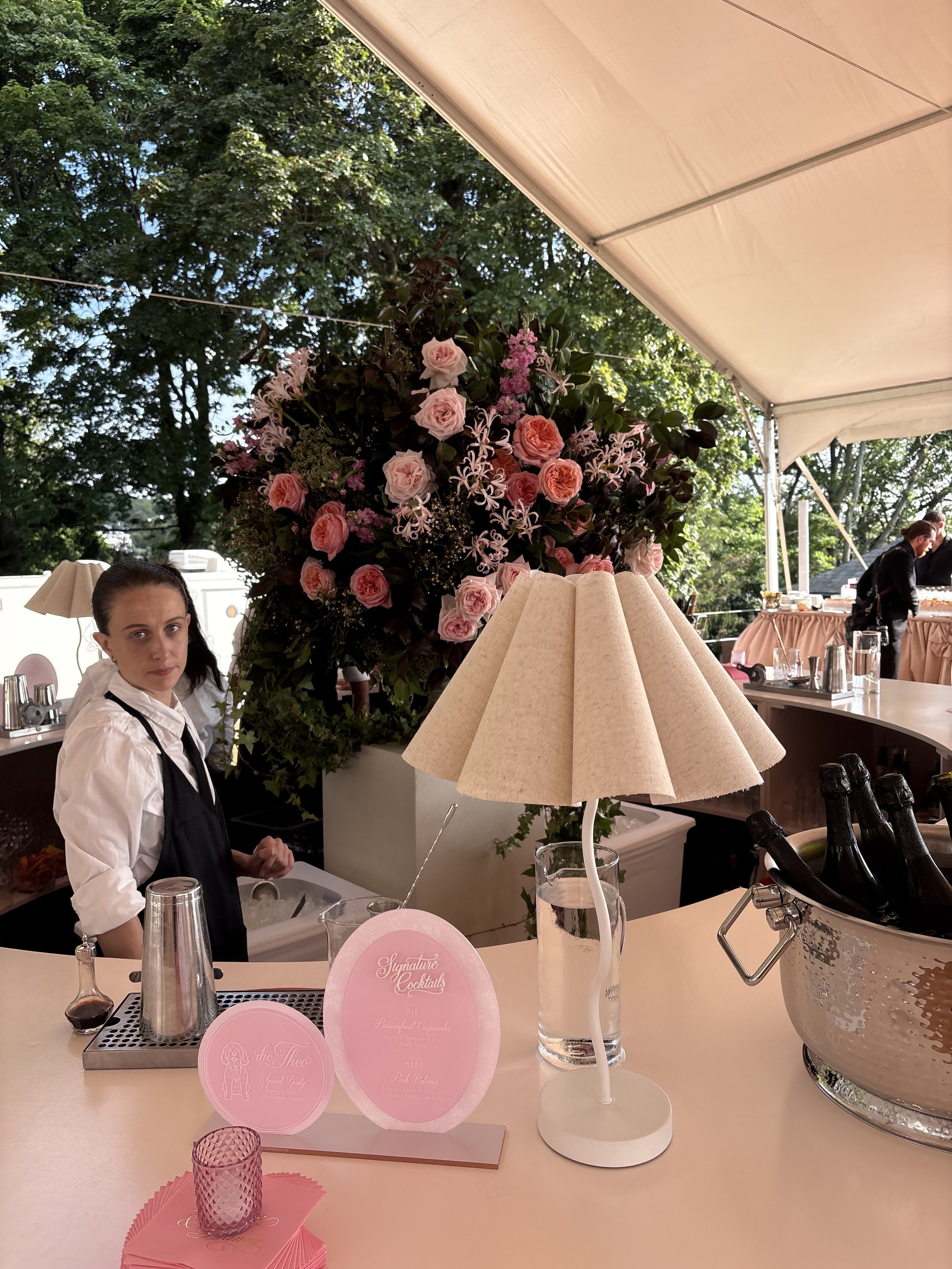A woman in a white shirt and black apron standing behind a bar counter with a pink flower arrangement, a table lamp, and various drink bottles. Outdoor setting with green trees and a beige canopy overhead.