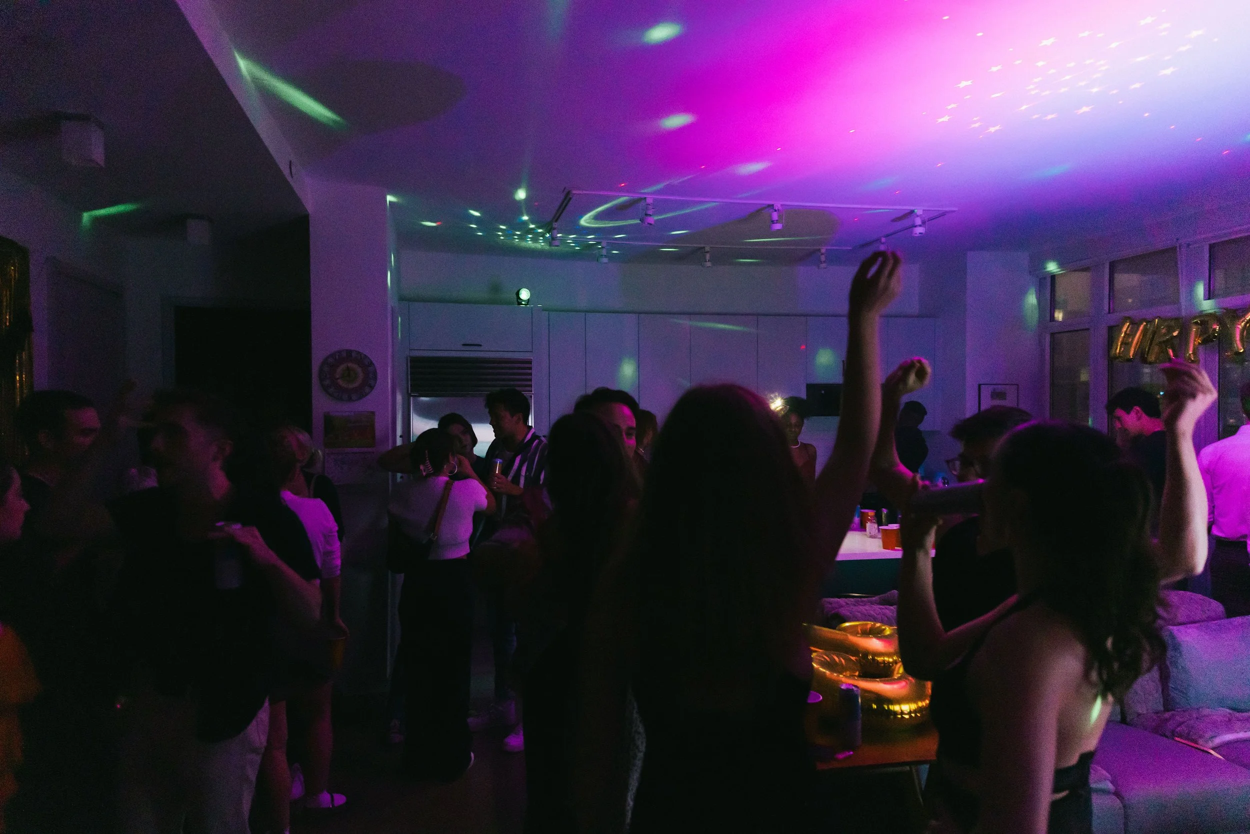 People dancing and socializing in a dimly lit room with purple and green laser lights projecting on the ceiling.