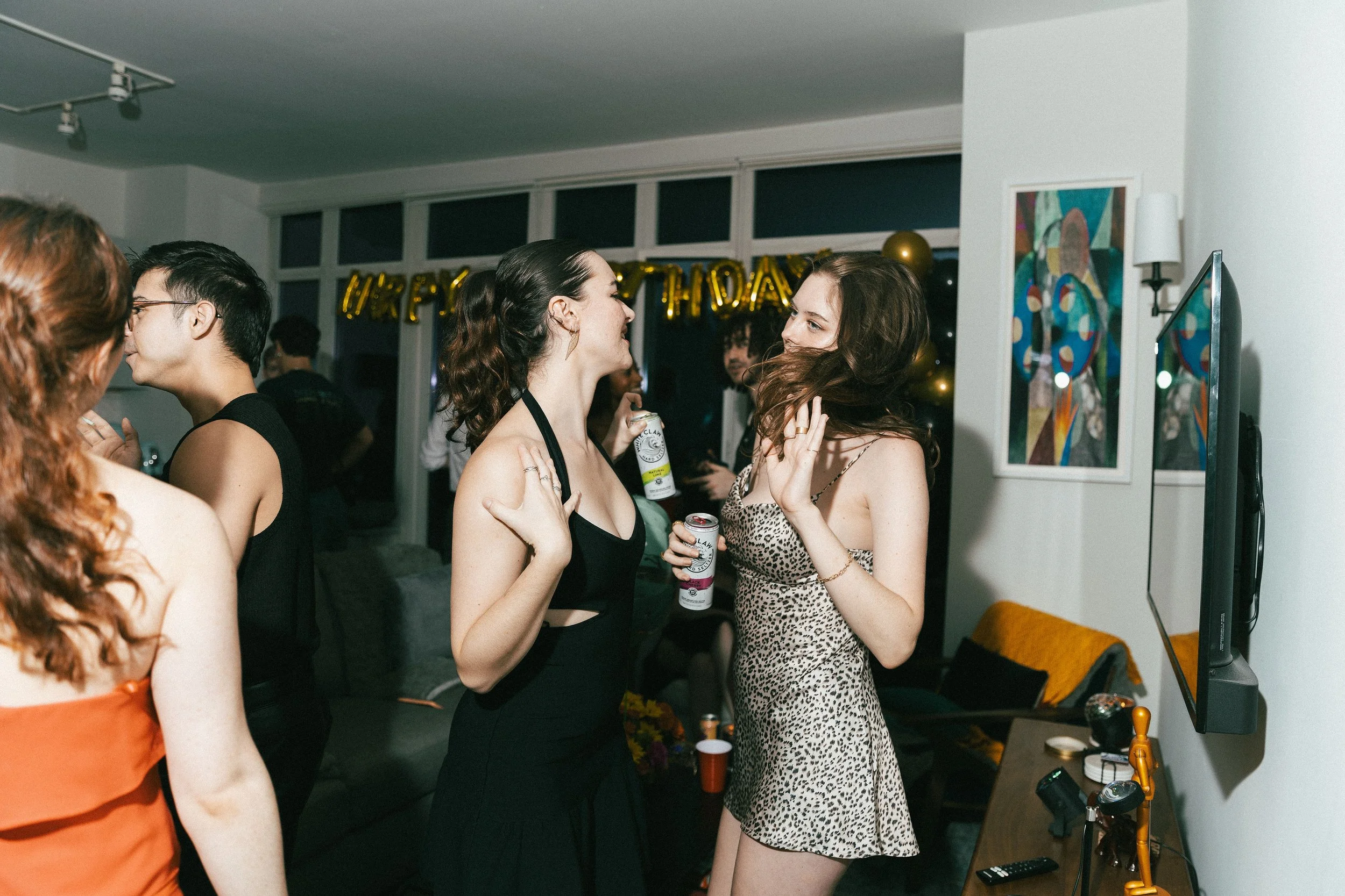 People celebrating at a party, some holding drinks, with a 'Happy Birthday' banner in the background.