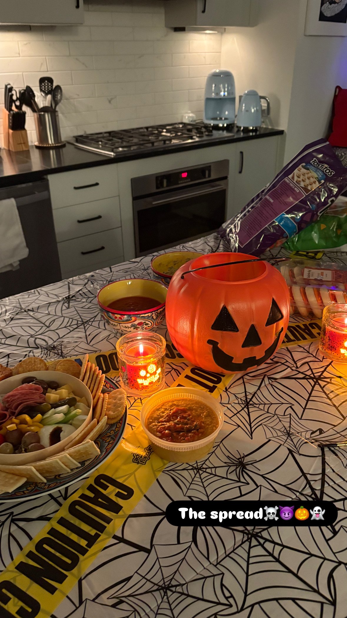 A Halloween-themed table setting with a carved pumpkin lantern, candles, snacks, and a red and black themed tablecloth featuring spider web patterns. The background shows a kitchen with a stove, kettle, toaster, and cabinets.