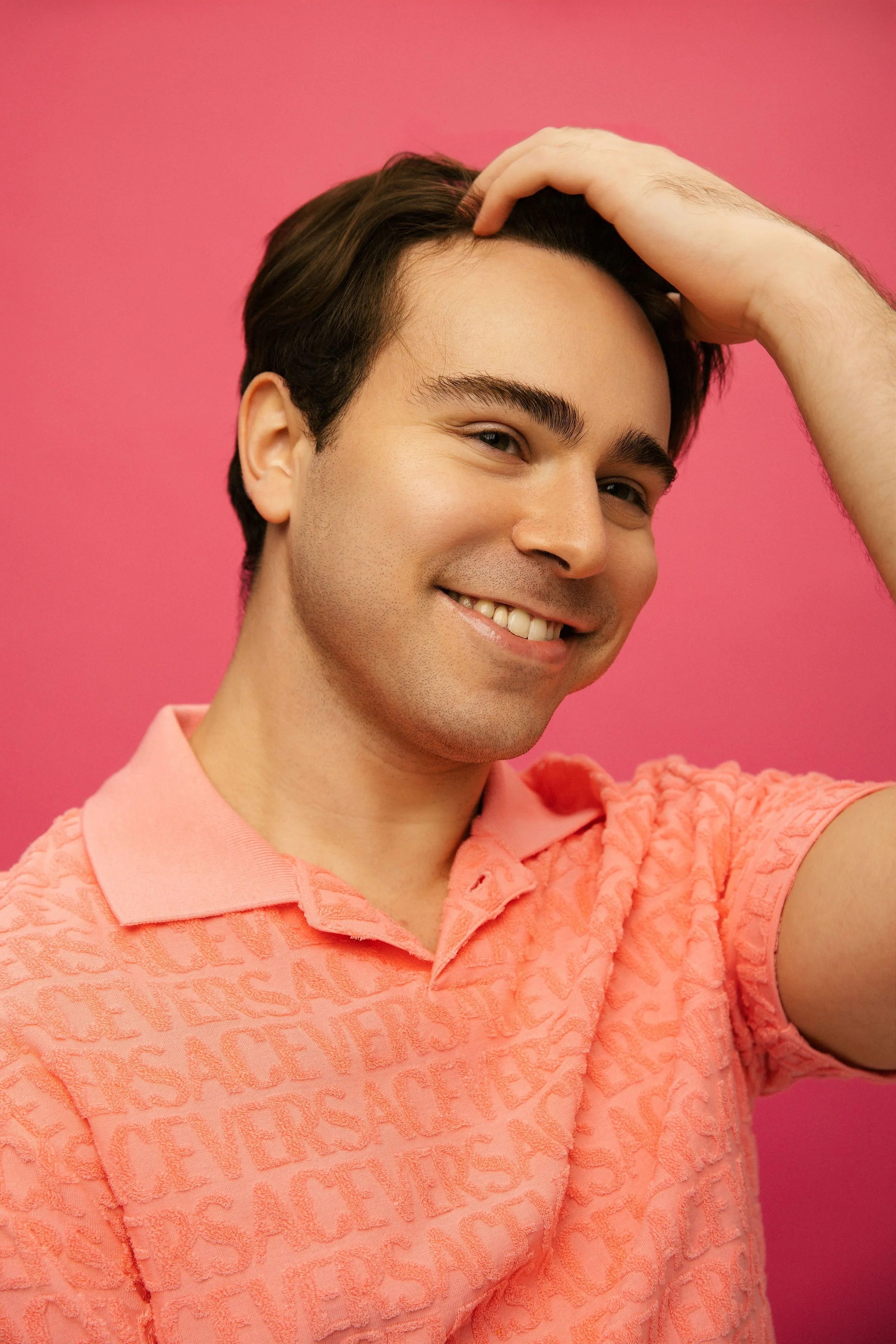 Close-up of a smiling man with dark hair and light skin, wearing a pink textured polo shirt, against a solid pink background.