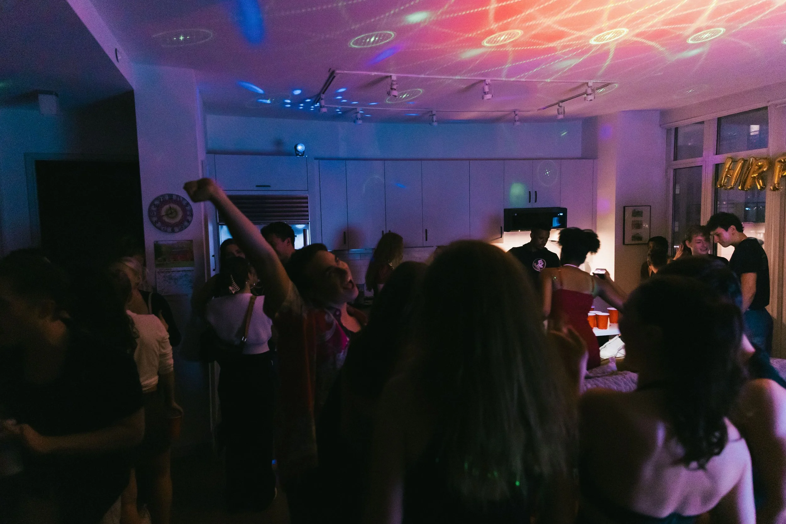 People gathered indoors at a party with colorful lighting and decorations, including gold balloons spelling 'HAPPY' near a window.
