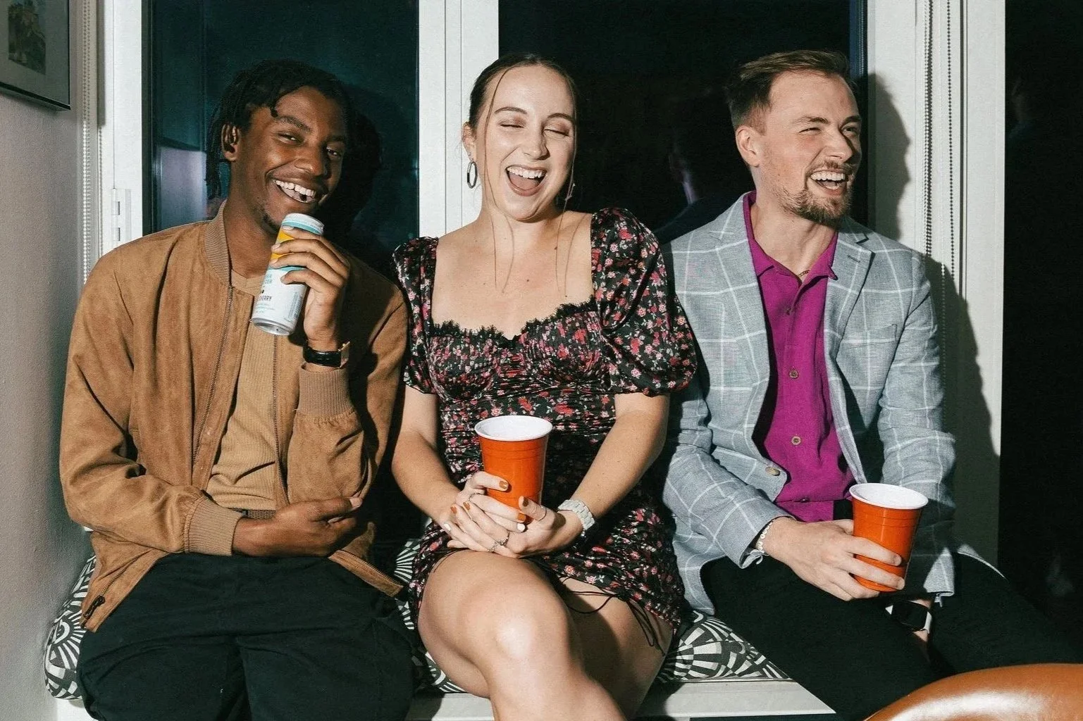 Three friends sitting together, laughing and enjoying drinks at a social gathering indoors. The person on the left is holding a can, the woman in the middle has a red cup, and the man on the right also has an orange cup.