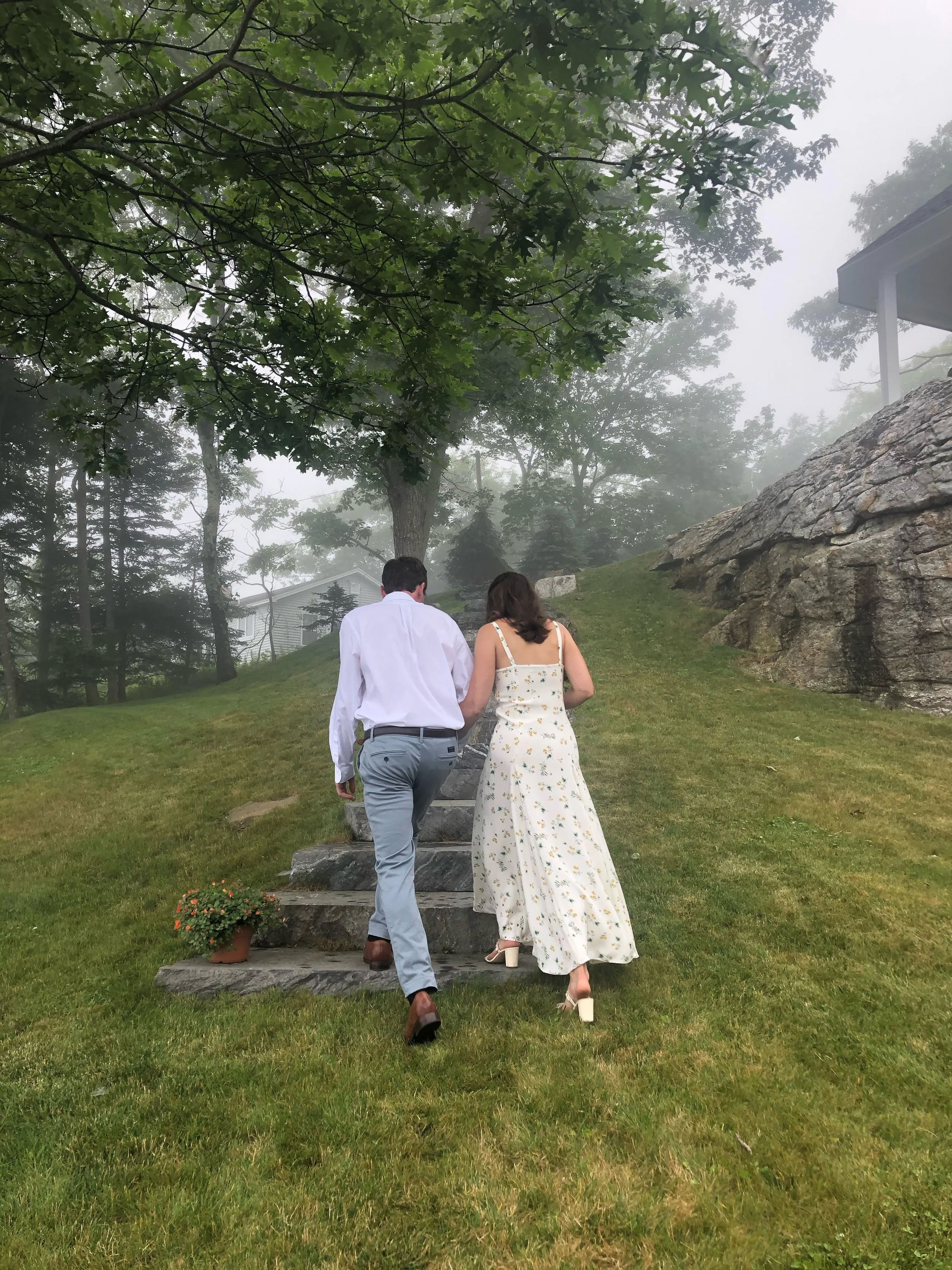 A couple walking up stone steps in a lush, foggy outdoor area. The woman is wearing a white dress with a floral pattern and high heels, while the man is dressed in a white shirt and light gray pants. The surroundings include green grass, large rocks, and numerous trees with dense foliage.