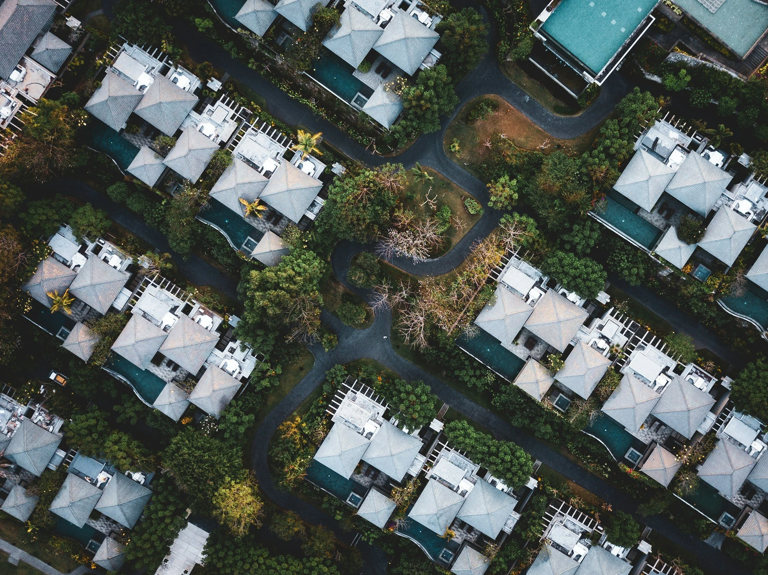 An aerial view of a residential area with houses, green trees, winding roads, and several swimming pools.