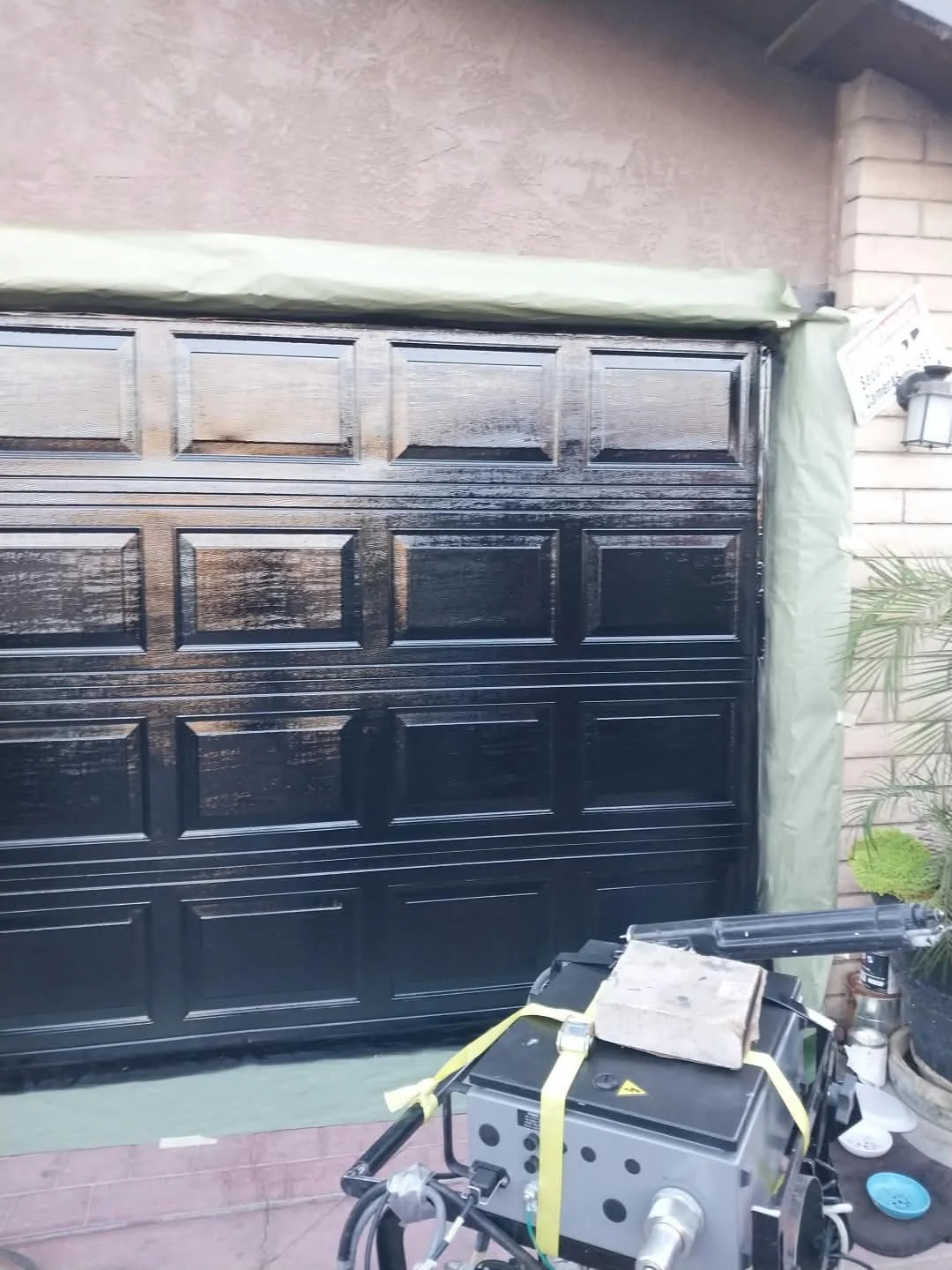 A black garage door being painted, with painting equipment and supplies in front, a tarp above, and a brick house with plants nearby.