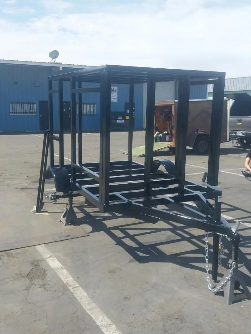 Empty black metal utility trailer with an open frame design, parked in a lot with a blue industrial building in the background.
