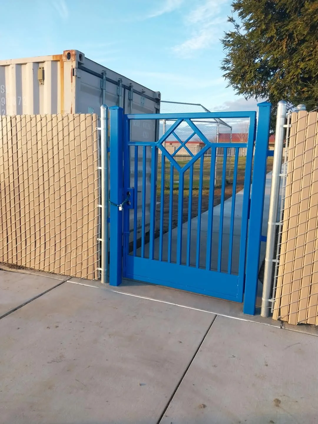 Blue metal gate with decorative diamond shape, gate lock, attached to beige chain-link fence, sidewalk, grassy area, large metal shipping container and trees in the background.