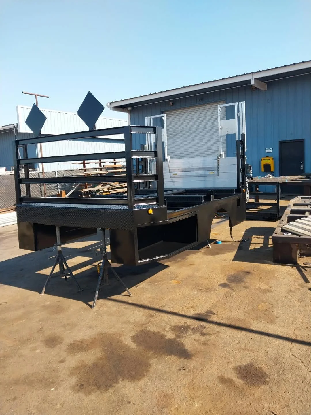 A flatbed truck with a black metal frame and railings on the sides, parked outside a blue industrial building. There are two diamond-shaped signs mounted on the front of the truck's frame and various tools and materials surrounding it.