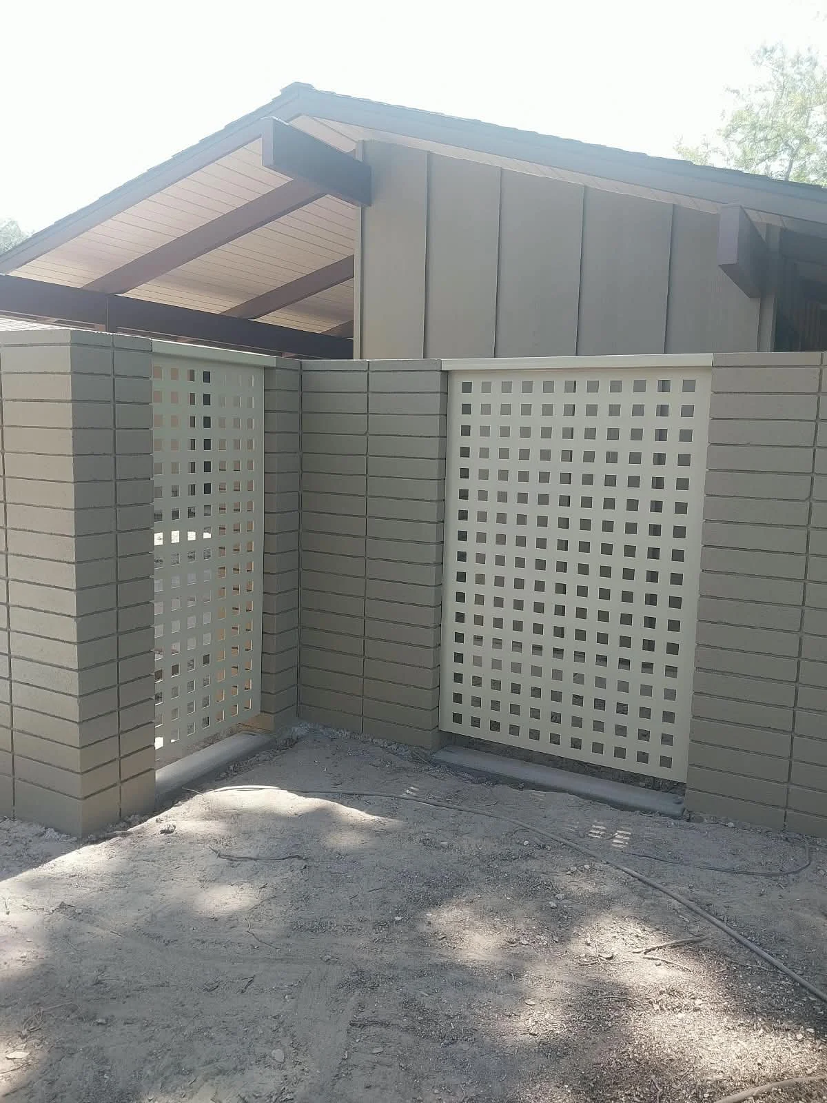 A beige lattice fence surrounds a house with a metal roof and exterior walls, with trees in the background and a dirt ground in the foreground.