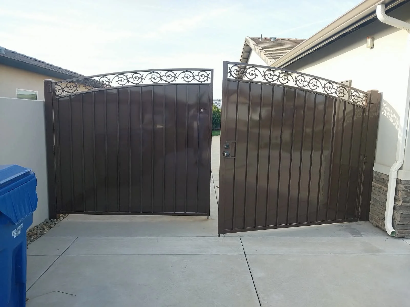 A black metal double gate with ornate scrollwork on top, partially open, leading to a driveway in front of a house with gray and white exterior, and a blue trash bin on the side.