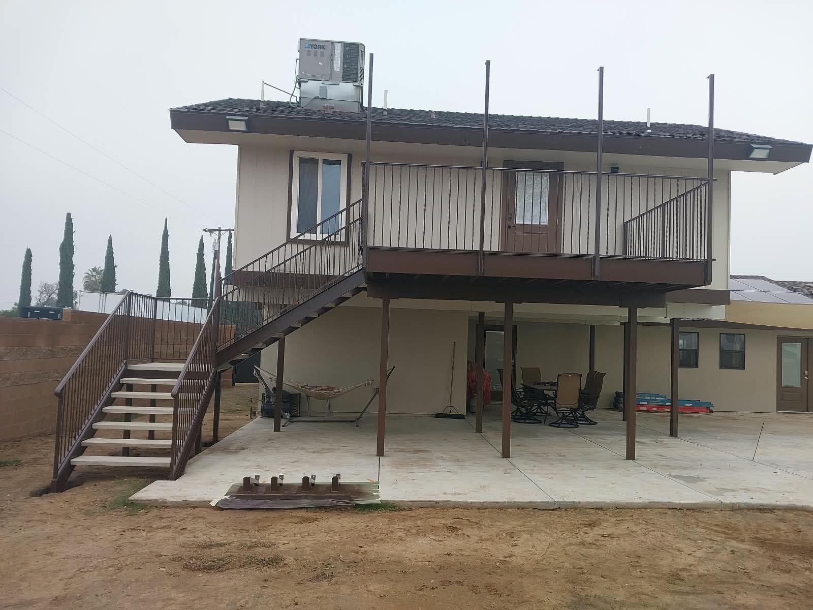 A two-story house with a stairway, balcony, and patio, with outdoor furniture and some construction or yard materials.