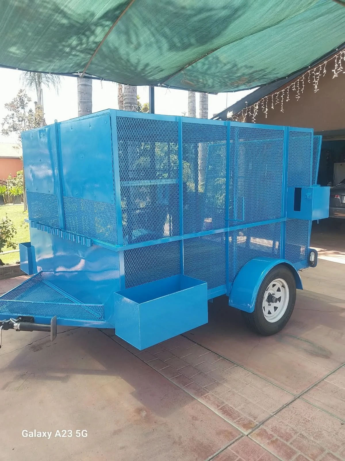 A blue mobile food or merchandise cart with mesh sides and a hitch for towing, parked under a shade cloth in a driveway, with trees and a house in the background.