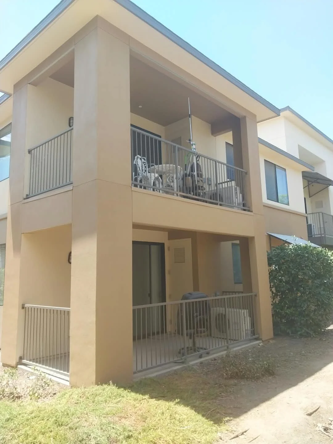 Two-story beige apartment building with balconies on both floors. The upper balcony has chairs, a table, and a scooter, while the lower balcony has a parked washing machine.