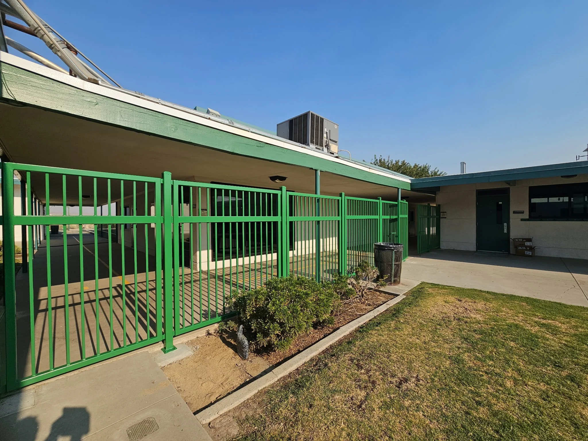 A school building with a green fence in front, a sidewalk, and a grassy area under a clear blue sky.