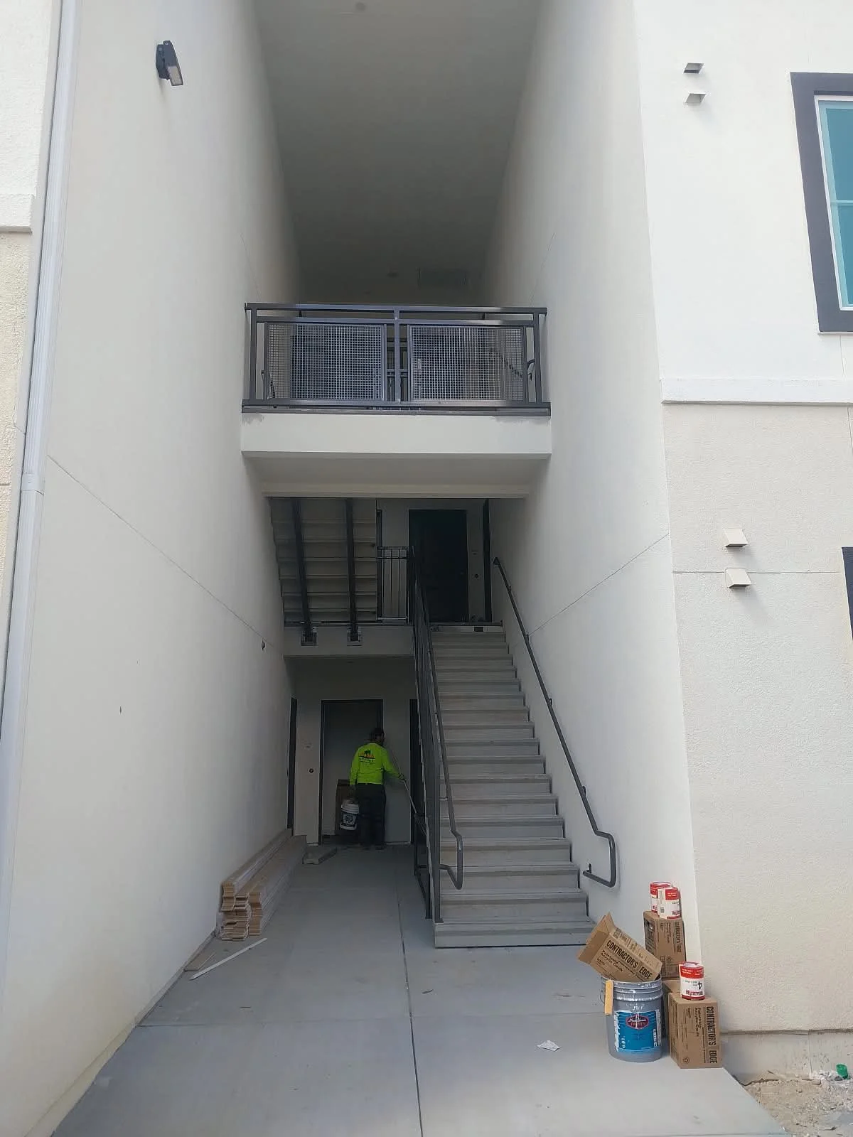 Exterior of a modern building with a staircase leading to the second floor, a worker in a neon yellow shirt working near the wall, and construction supplies on the ground.
