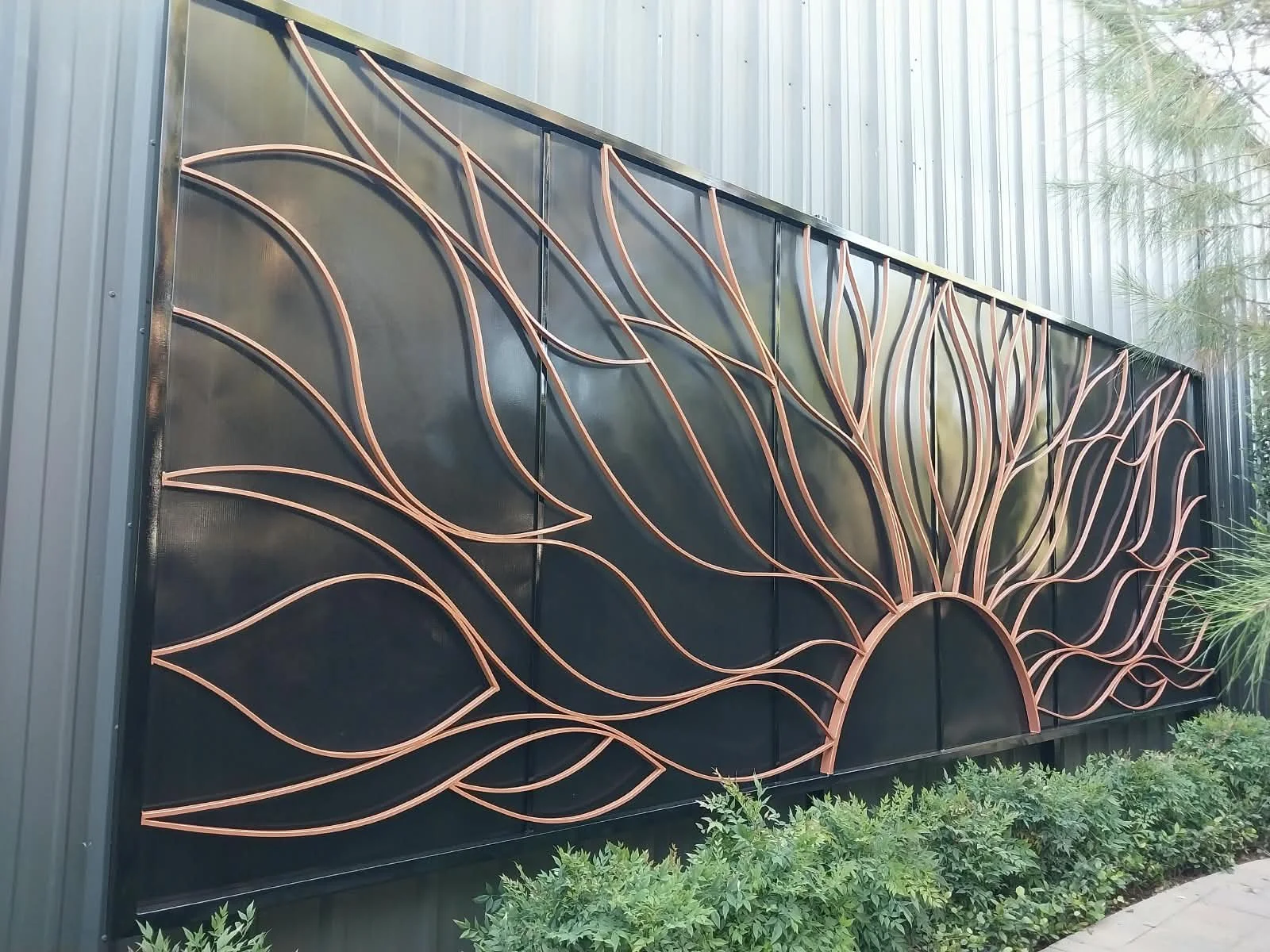 Decorative black metal panel with a rose gold abstract design resembling a stylized sun and floral pattern, mounted on a building exterior with nearby greenery.