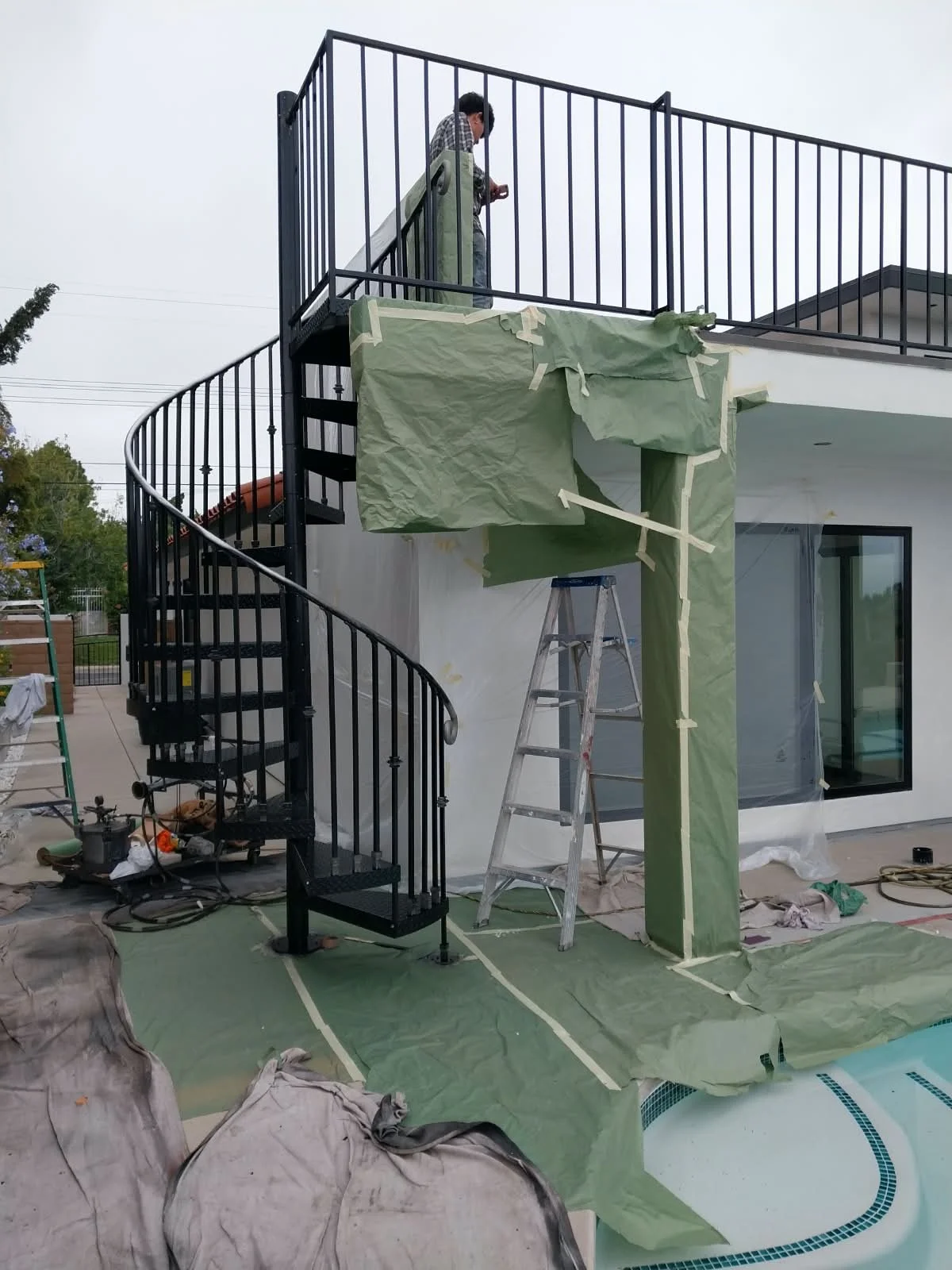 Person on ladder doing construction work on an outdoor curved staircase and balcony of a modern house, with protective green paper and painter's tape.
