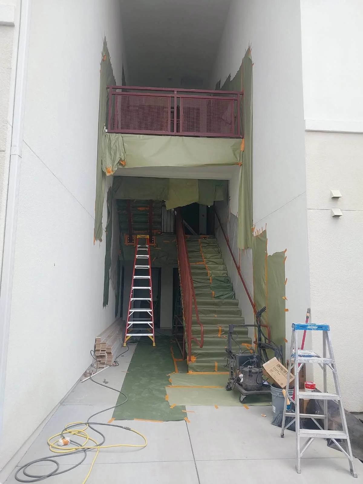 Construction site with staircase and scaffolding, green protective covers, ladders, and building materials, inside a building with white walls.